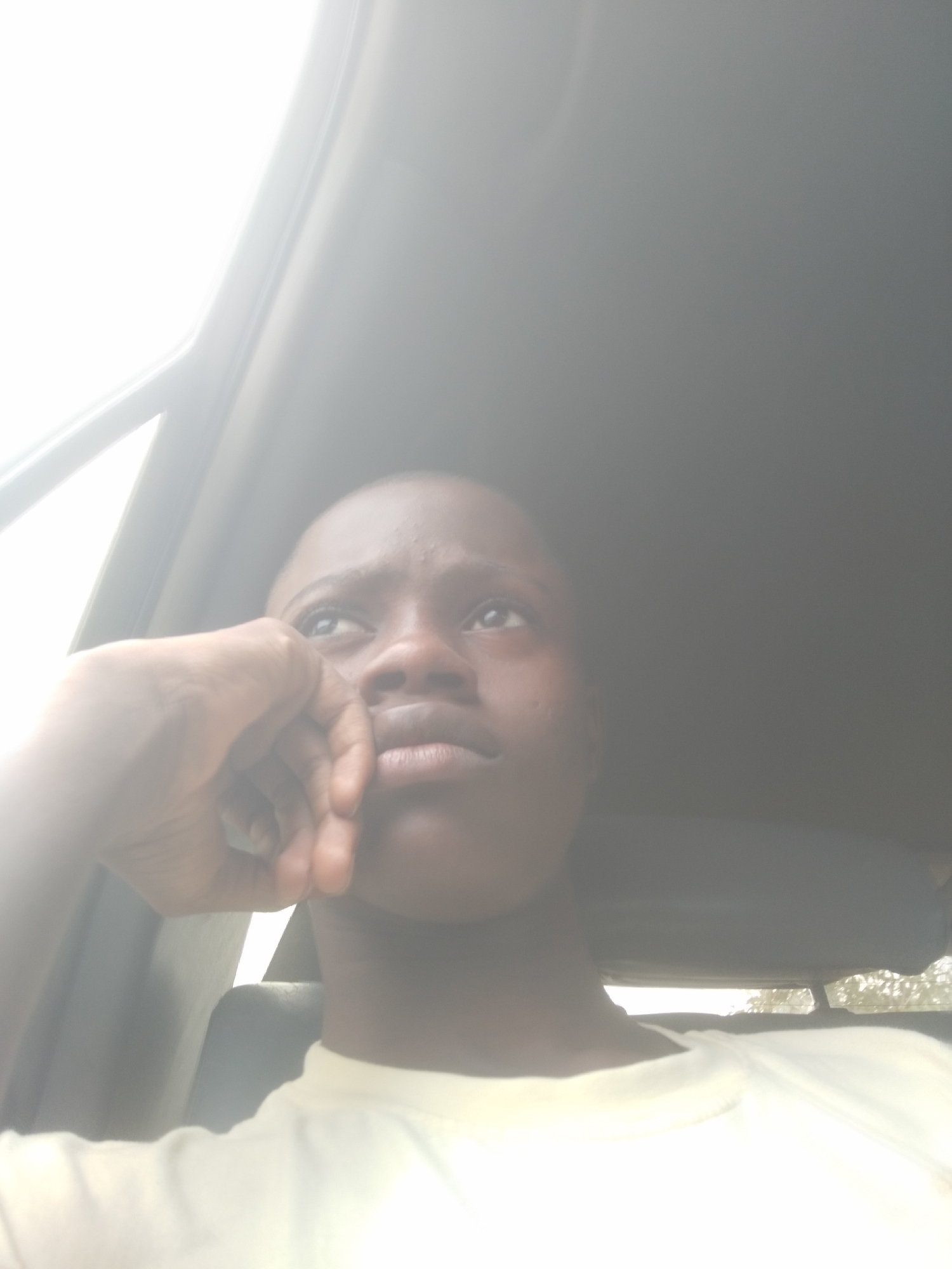 A young person with dark skin is sitting in a car. The person is wearing a light-colored t-shirt and resting their head on their hand, looking upwards. The location appears to be a city as indicated by the background trees, but the specific city is Unknown, Unknown.