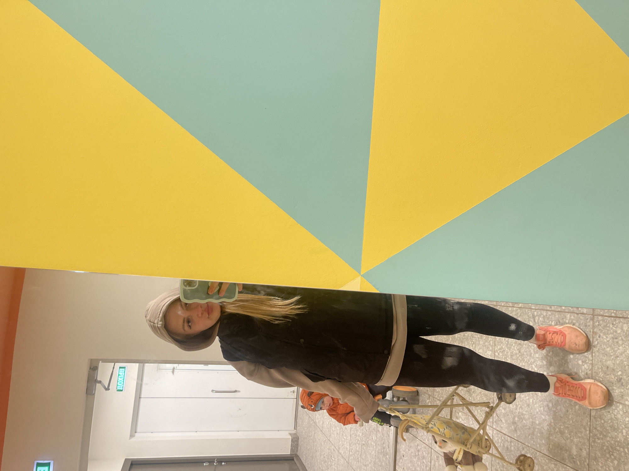 A young woman in a tan hoodie, black jacket, and black leggings stands in what appears to be a hallway, taking a mirror selfie with her smartphone. She has blonde hair and is wearing pink athletic shoes. Behind her and visible in the mirror is a section of a wall painted with intersecting yellow and teal geometric shapes. Also reflected in the mirror are an exit door and an orange jacket with a fox hood, with a stroller. The location is Izhevsk, Russia.