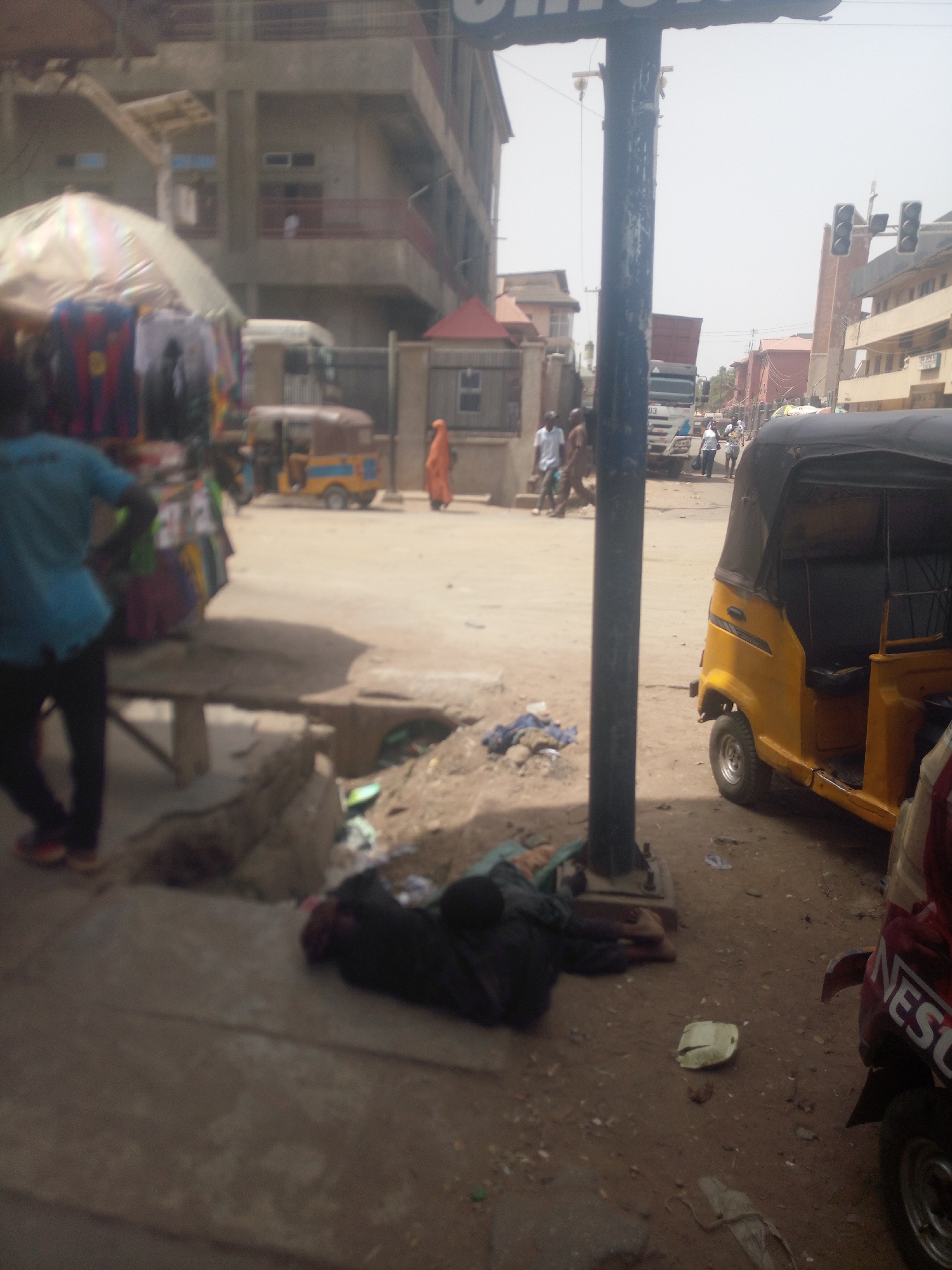 The image depicts an urban scene in Maiburiji, Nigeria. A person is lying on the sidewalk next to a light pole. Debris litters the ground. A yellow three-wheeled vehicle (auto rickshaw) is parked nearby. Further down the street, people are walking, and other auto rickshaws and a truck are visible. Traffic lights are in the background. A clothing stall sits on the left.