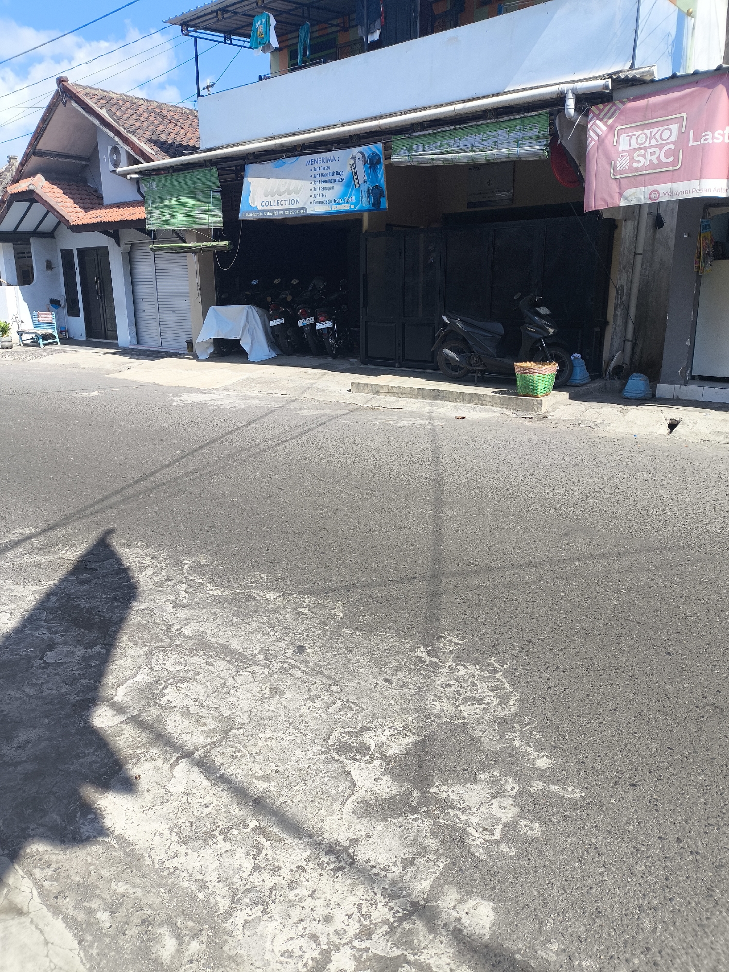 This is a photograph of a street scene in the city of Citrodiwangsan, Indonesia. The image shows a commercial building with a street in the foreground. The building appears to be a shop or business, with a dark-colored gate and a sign that says "TOKO SRC Last." There are several motorcycles parked under an awning in front of the building. To the left of the building, there is a residential house with a tiled roof. The street is paved with asphalt and shows signs of wear and tear, with some patches of lighter-colored concrete or repairs. The sky is blue with some white clouds. A shadow of a person is cast on the street in the foreground.
