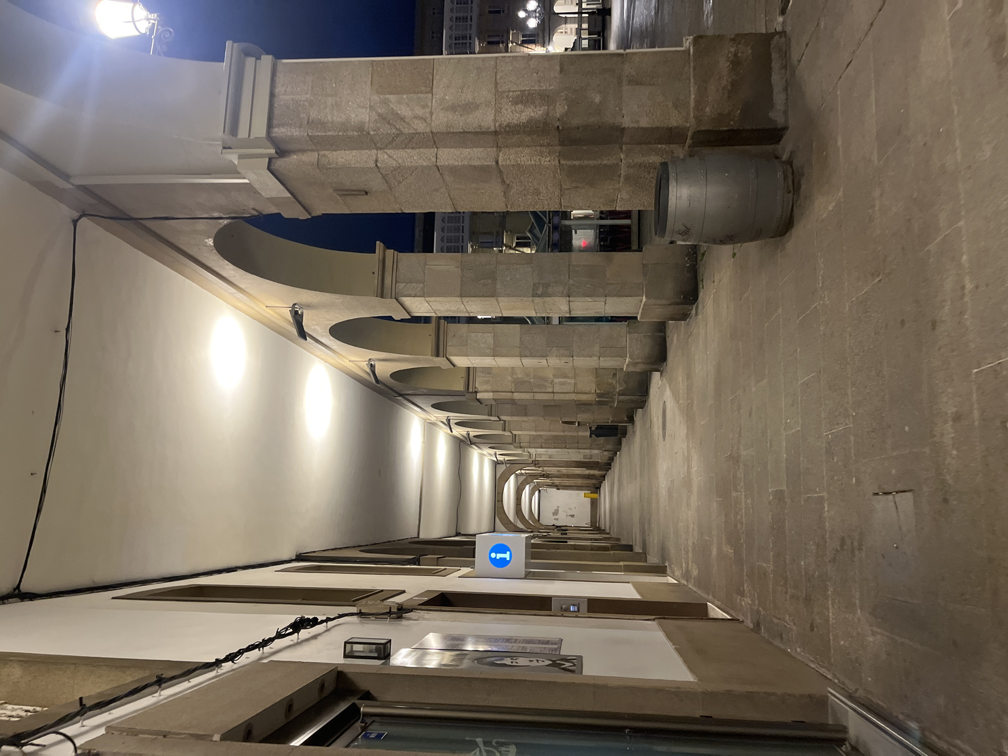 The image depicts a long, covered walkway in A Coruña, Spain, viewed from a low angle, looking towards the far end. The walkway is defined by a series of stone arches on the right side, supported by pillars made of stacked stone blocks. A large, metal barrel is attached to one of the pillars on the right. The left side of the walkway features a white wall with recessed rectangular spaces near the top, below the arches. Below the recessed spaces are shopfronts. Near the foreground, a bright blue cube with a white "i" symbol is visible. The lighting is dim, with bright circular lights illuminating the left wall beneath the arches. Streetlights are visible in the distance at the end of the walkway. The ground is paved with stone. The image has been rotated by 90 degrees.
