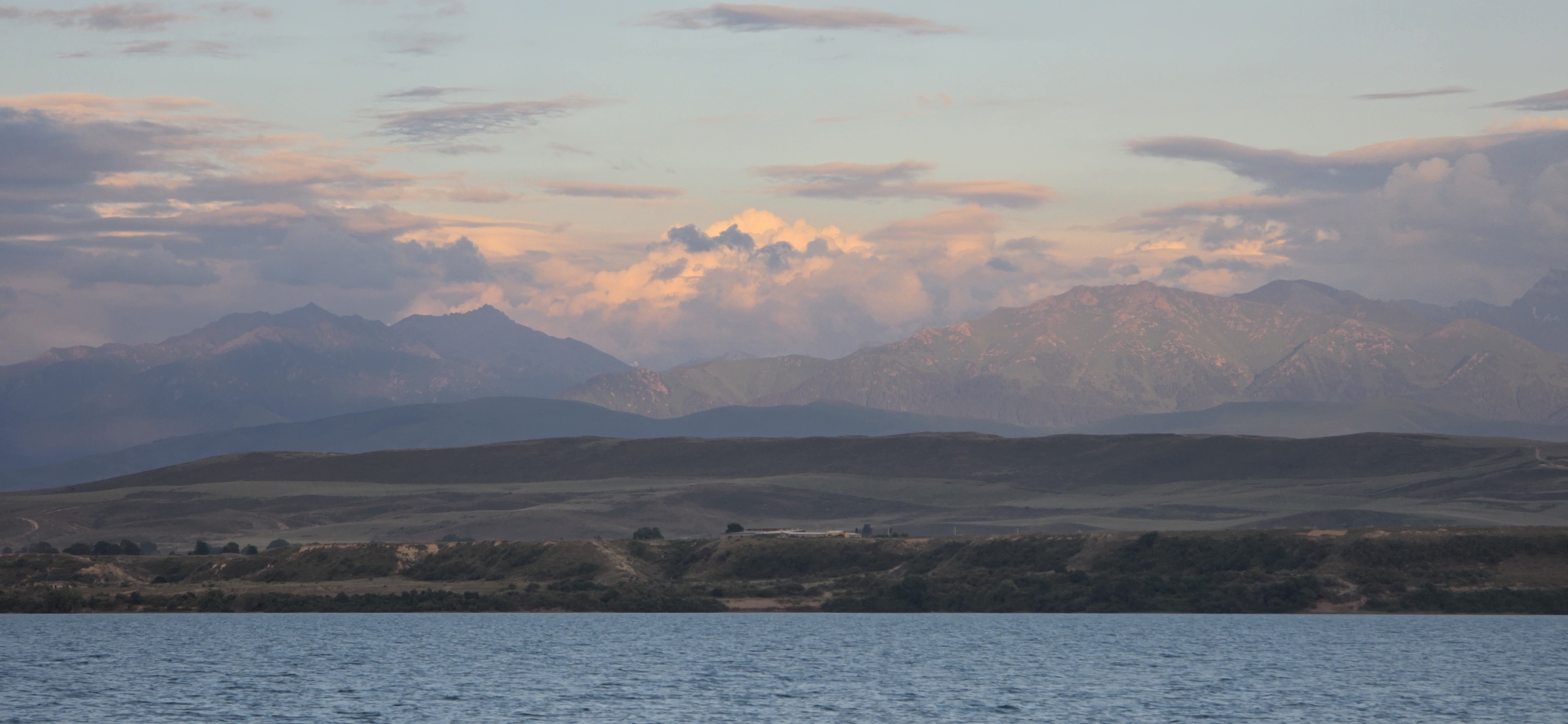 A wide, horizontal shot captures a tranquil scene of a lake in the foreground, leading to rolling hills and majestic mountains in the background. The sky above is a canvas of pastel hues, with soft pink and orange clouds scattered across a pale blue expanse. The mountains, tinged with the warm glow of sunset, are layered in varying shades of purple and blue, creating a sense of depth. The water in the lake is a muted blue, with subtle ripples disturbing its surface. The overall impression is one of serene natural beauty, possibly at dawn or dusk, in a mountainous region. The location is identified as Pristan'-Przheval'sk, Kyrgyzstan, a city situated on the eastern shore of Lake Issyk-Kul.
