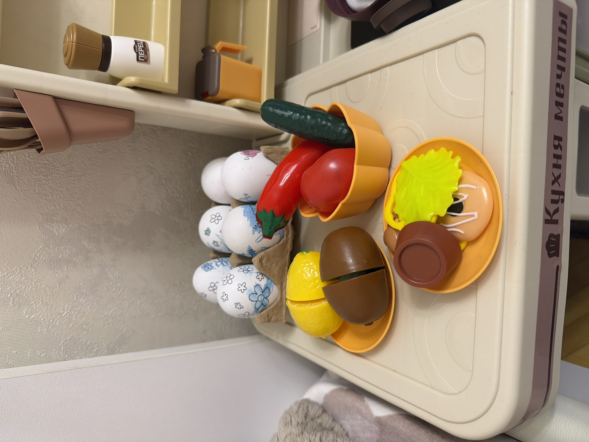 In an indoor setting, a toy kitchen playset is depicted. The play kitchen has a beige and white color scheme. On the top shelf of the playset, a white pepper shaker with a brown cap is visible, along with a small orange bucket. Below this, a carton holds four toy eggs, each decorated with blue floral patterns on a white background. In front of the egg carton, a yellow toy lemon, sliced in half, rests on an orange plate. Next to the lemon is a toy kiwi, also sliced in half, on the same orange plate. To the right of the lemon and kiwi, an orange bowl contains toy vegetables: a red chili pepper, a red tomato, and a green cucumber. Below these vegetables, another orange plate holds a toy donut with white icing and a green toy lettuce leaf. The playset has a label at the bottom with text in Russian, which translates to "Kitchen of Dreams." The background consists of a wall with textured wallpaper in a neutral tone. The overall scene appears to be a collection of toy food and kitchen accessories.