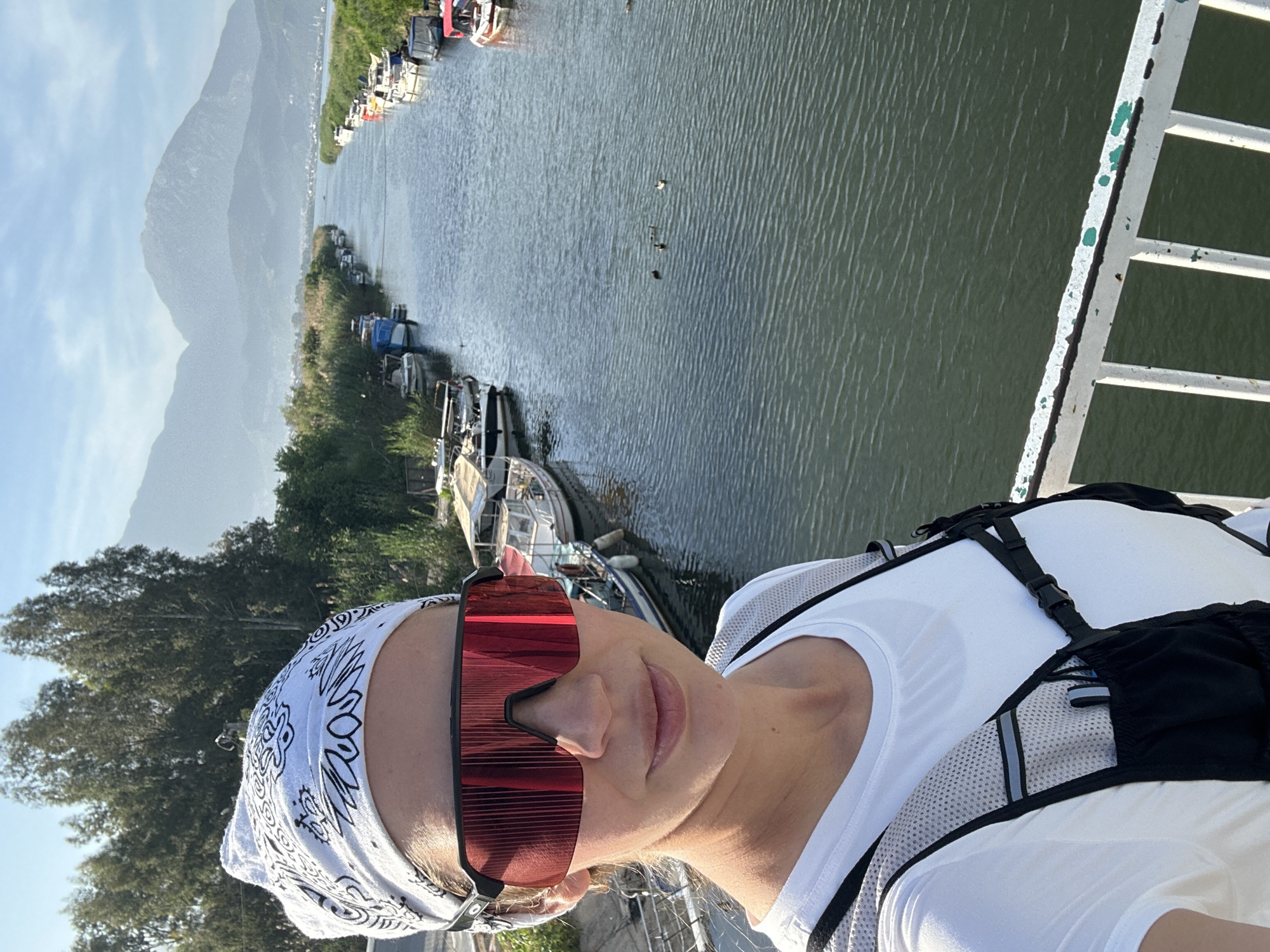 The image shows a person, likely female, wearing sunglasses and a bandana, possibly taking a selfie. She is wearing a white shirt and a backpack. The background features a river with boats docked along the banks. In the distance, there are mountains. The photo appears to be taken from a bridge. This location is likely in Fethiye, Turkey due to the presence of the river and mountains which are commonly found in the area.