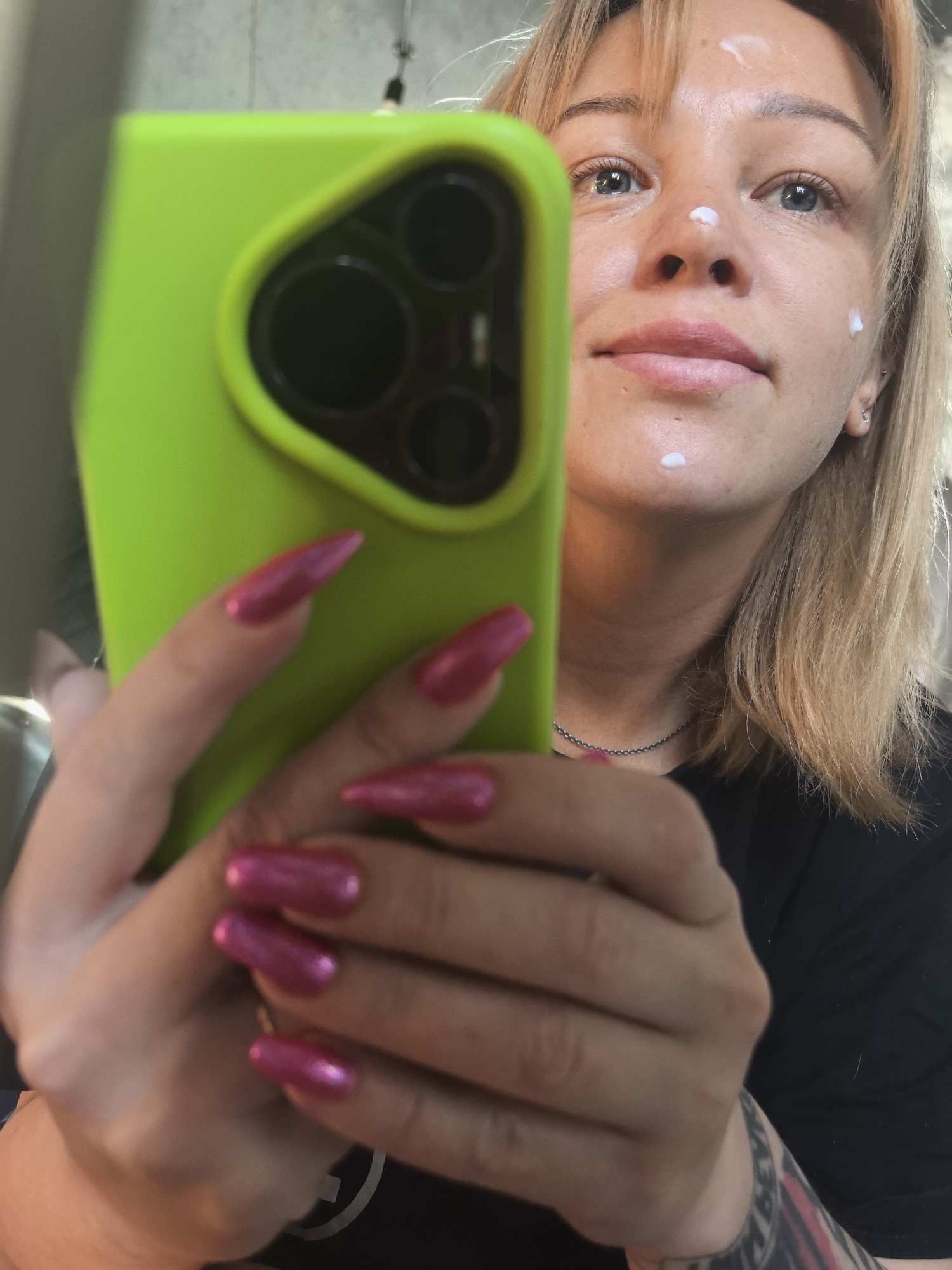 A woman is taking a selfie with a green smartphone, focusing on her face, which has dots of white cream on her nose, cheek, and forehead. She has blonde hair with bangs, blue eyes, and a silver necklace. Her left hand holds the phone, showcasing long, pink, pointed nails. A tattoo is visible on her lower arm. The background is blurred. There is no specific information about the location being Noviy, Russia.