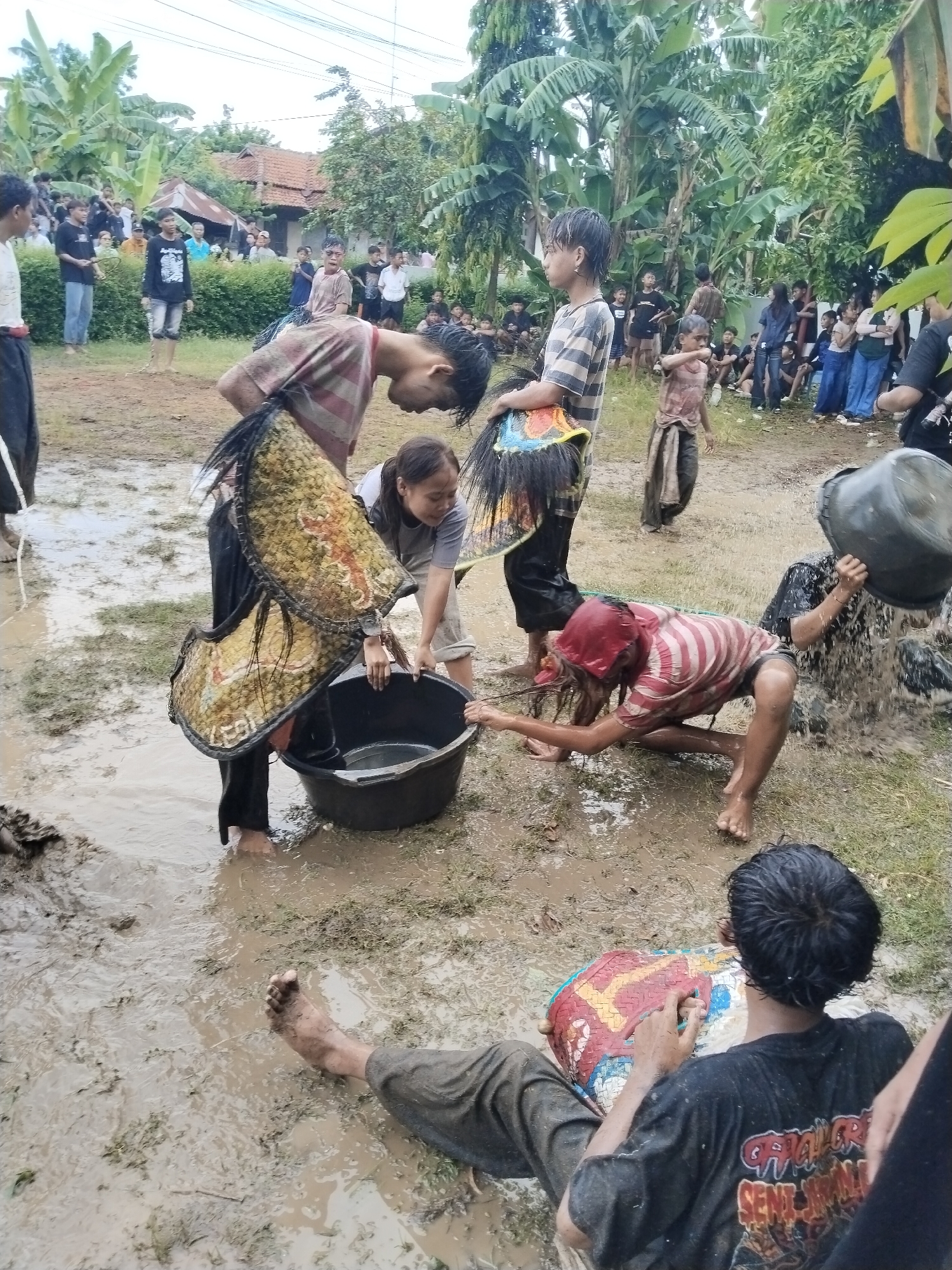 In a muddy field in Karang Tengah, Indonesia, a group of people, mostly young, are interacting with water. Some individuals are pouring water from buckets onto others who are squatting or kneeling in the mud. Several people are standing around the perimeter observing. Two carry what appear to be ceremonial horse effigies. The environment is rural, with houses and trees in the background.
