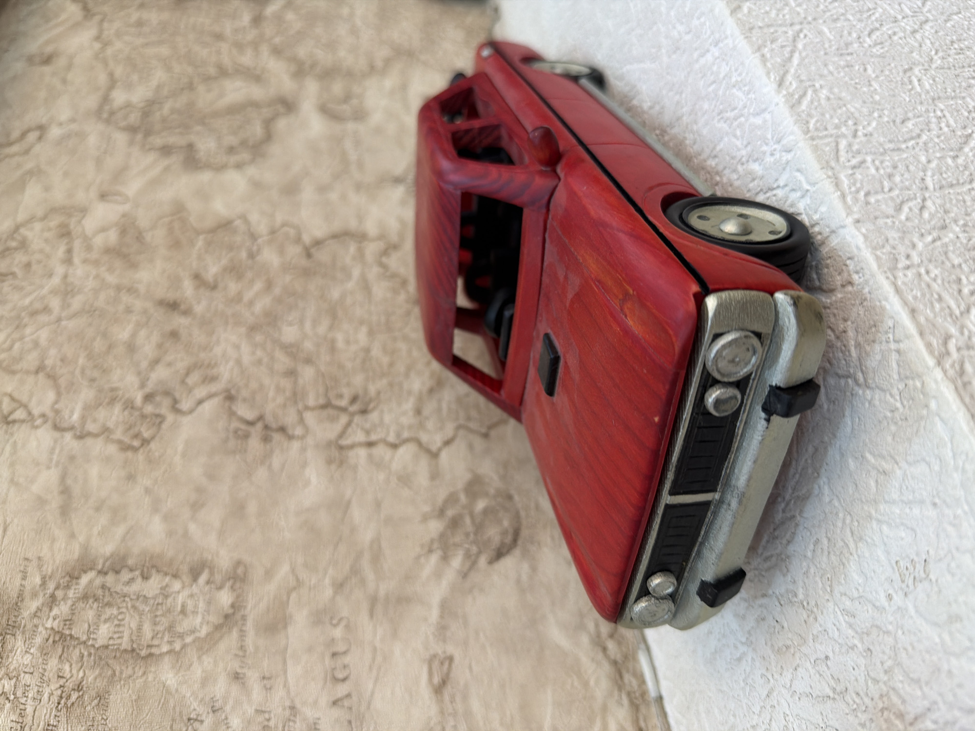 An overhead shot shows a red toy car model with a silver bumper and black trim parked on a textured white surface. The car is positioned diagonally, facing left. The background features a faded, sepia-toned map with illegible text and geographical features. There is no visible indication of the city of Sabirabad, Azerbaijan.