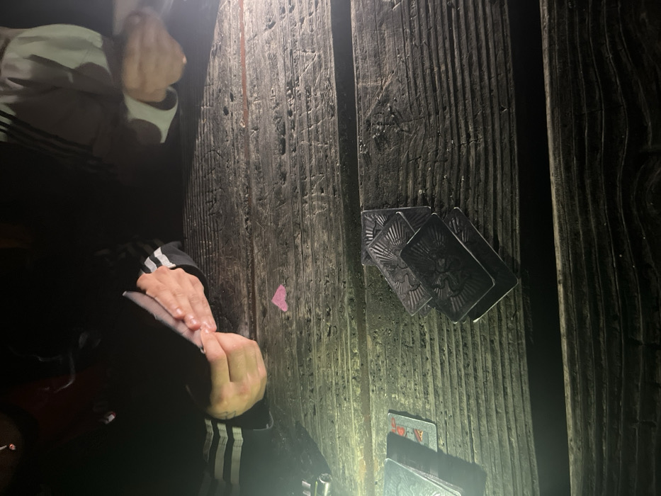 This image captures a low-light, outdoor scene in Strasbourg, France, likely at a public park or similar outdoor setting with a picnic table during nighttime. A strong, direct light source, possibly a flashlight or phone light, illuminates the weathered, dark wooden table from the lower left, casting shadows.
At least two individuals are present, visible primarily by their hands and forearms. On the left, a person wearing a dark long-sleeved garment with three white stripes is seen manipulating a small, unidentifiable object with both hands. A metallic, silver-colored object is partially visible on the table to their far left. Near the center of the table, in front of this individual, rests a small, single pink heart-shaped object.
On the right side of the table, another person's hands are visible, also wearing a long-sleeved garment, possibly light grey with darker stripes, engaged with the table's contents. Multiple stacks of playing cards are scattered across the table. A prominent stack of four or five cards with dark, intricately designed backs (featuring what appears to be a stylized creature or emblem) is located on the lower right side of the table. To the far left, a separate, smaller stack of cards includes at least one card partially visible with a red suit symbol (possibly a heart or diamond) and a face card design. The overall scene suggests an ongoing card game or social gathering.