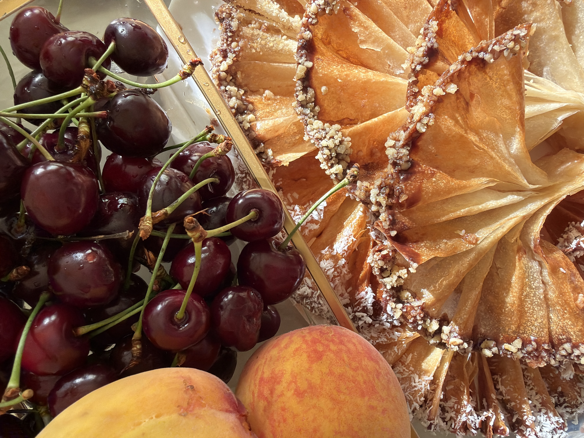 The image is a close-up, overhead shot of a collection of fruits and pastries. On the left side of the frame, a cluster of dark red cherries with green stems is prominently displayed. Some of the cherries have small droplets of water on them, suggesting they may have been recently washed. In the lower part of the image, two peaches with mottled orange and red skin are partially visible.
To the right of the fruits, a golden pastry is arranged in a circular, fan-like shape. The pastry appears to be made of thin, flaky layers, possibly phyllo dough, and is topped with chopped nuts and shredded coconut. A thin layer of syrup or glaze also seems to be present, giving the pastry a slightly glossy appearance. The pastry is decorated with a sprinkle of white shredded coconut along its edges. A golden-colored element, possibly a border or part of the serving dish, runs vertically between the cherries and the pastry.
The lighting suggests it is daytime, with bright natural light illuminating the scene. The background is indistinct, but the overall impression is that of a fresh and appealing presentation of food, possibly a dessert or a snack. There is no text visible in the image.