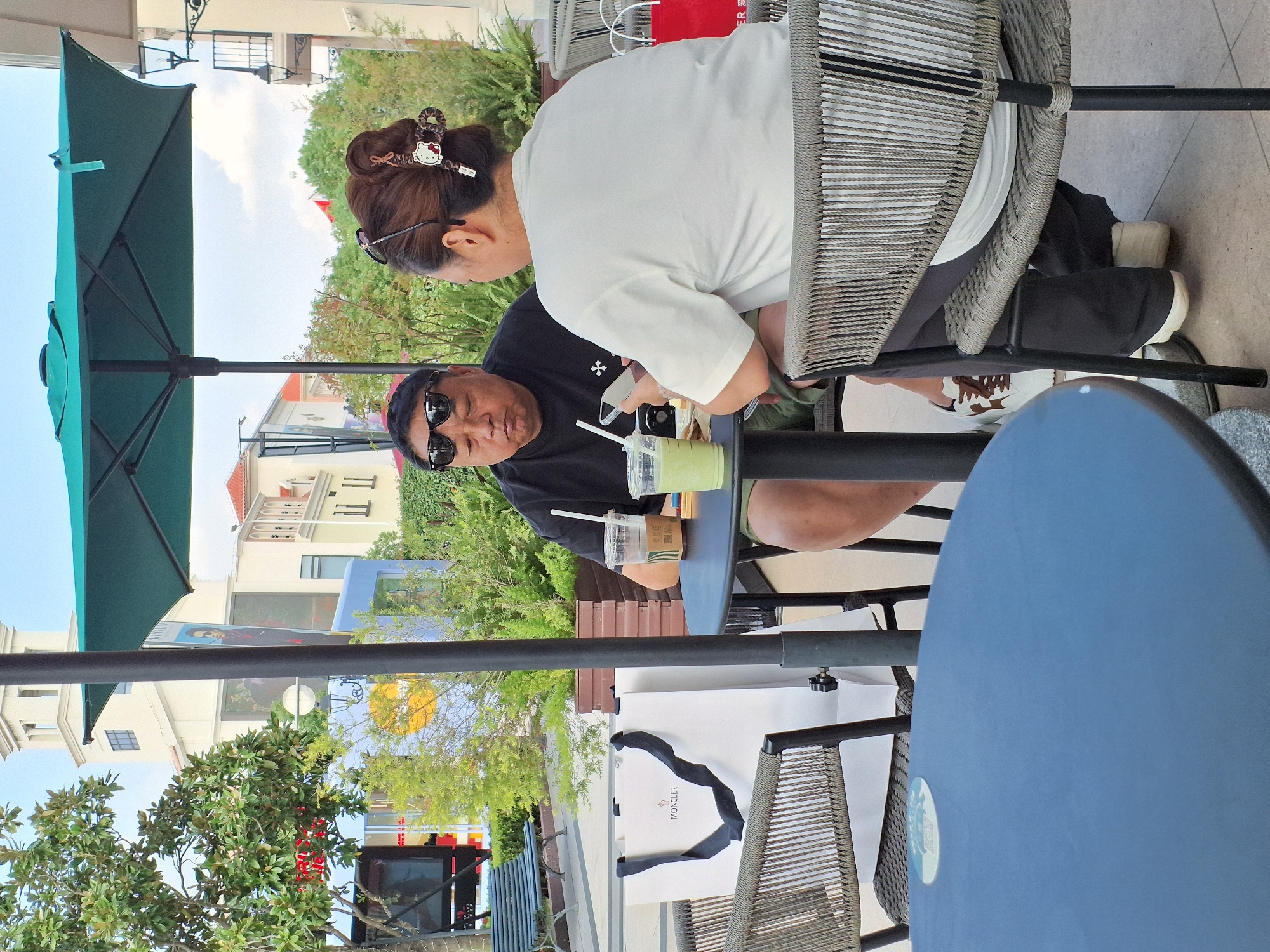 In Pudong, China, a man and a woman are seated at a dark round table outside a cafe, under a teal umbrella. The man, wearing sunglasses and a black t-shirt, is looking down at his phone. The woman, with her hair styled up and a decorative clip, is also looking at her phone. Two drinks, one in a clear cup with a straw and another in a similar cup, are on the table, along with a white shopping bag bearing the "MONCLER" logo. The setting appears to be an outdoor shopping area with buildings in the background and lush green foliage.