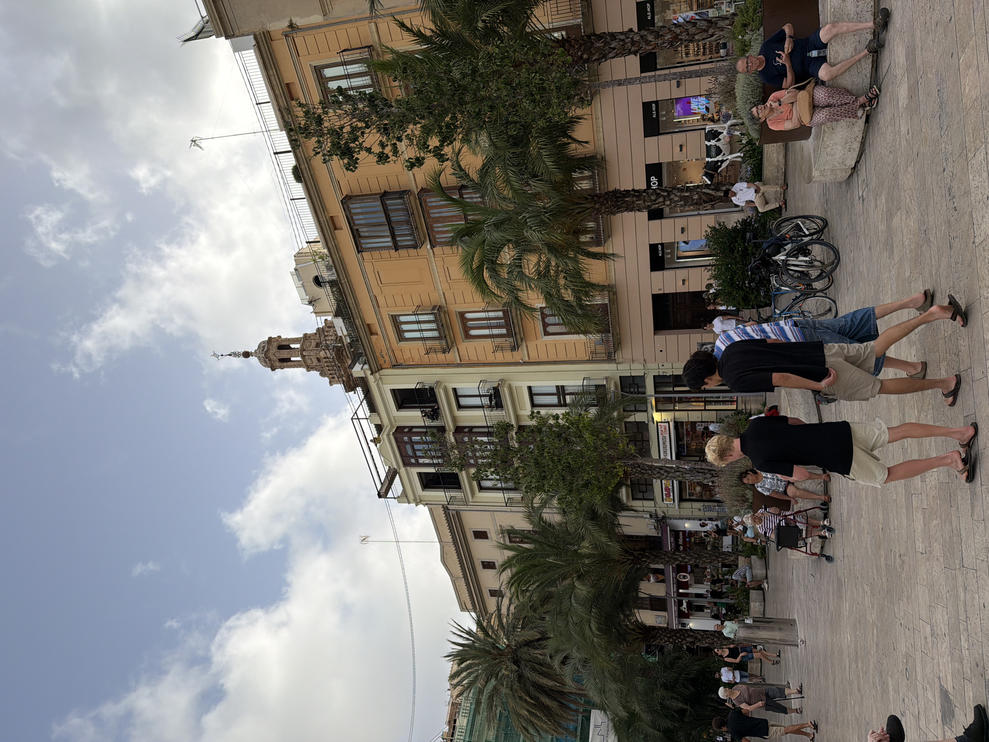 The image shows a street scene in a European city with a mix of old and new architecture. In the foreground, several people are walking and sitting. Two young men in casual clothing are walking away from the camera, one wearing a black t-shirt and khaki shorts, the other a striped t-shirt and denim shorts. Further back, an elderly woman with a walker is seated on a bench, and a couple is sitting next to her. Several palm trees are scattered throughout the scene. In the background, there are multi-story buildings with balconies and windows, one of which has a tower with a cross on top. Storefronts are visible at the ground level of some buildings. The sky is partly cloudy.