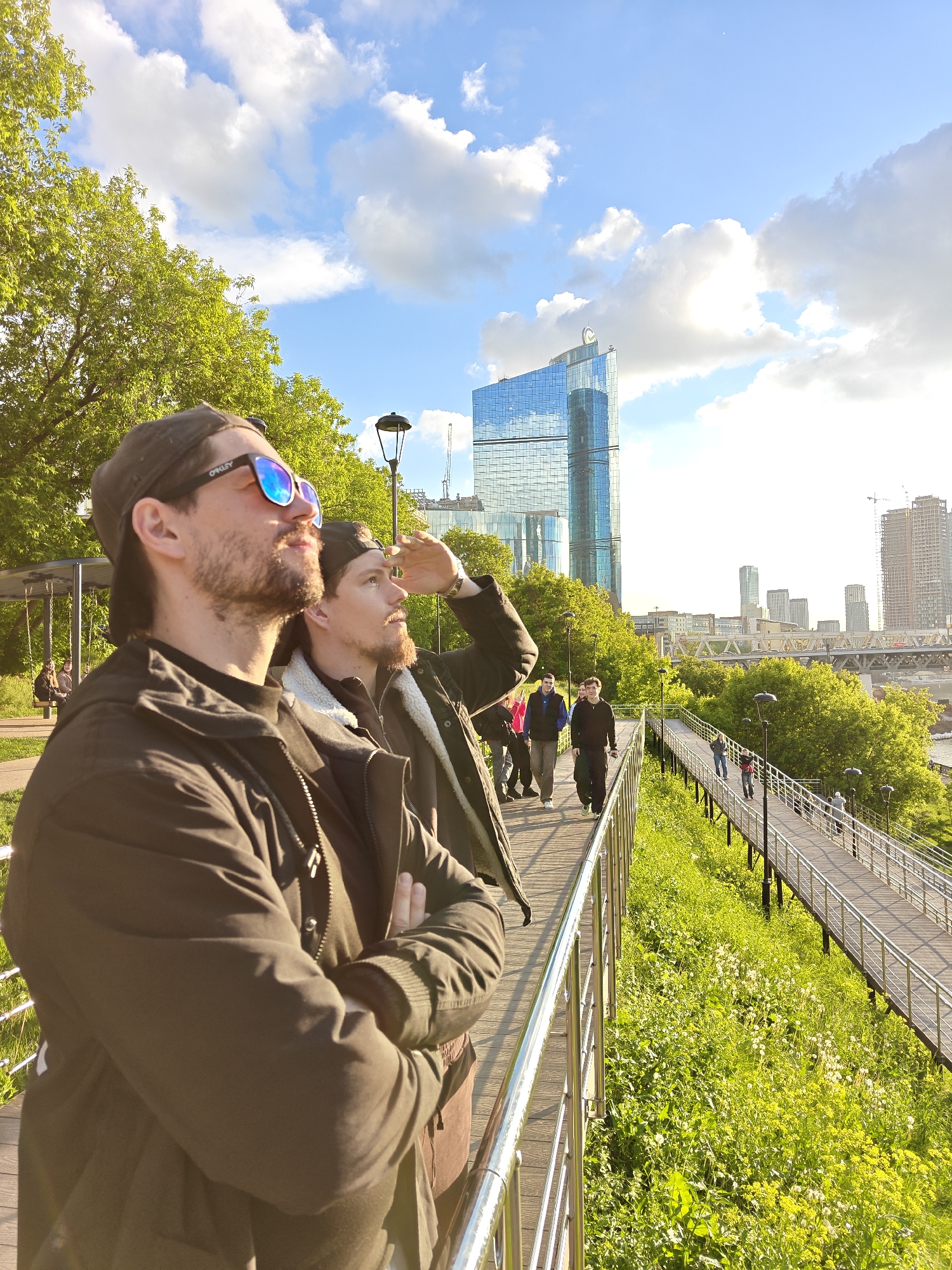 Two men stand at a railing, looking up. The man on the left wears sunglasses and a baseball cap. The man on the right has his hand shading his eyes. Behind them, a pedestrian walkway leads toward buildings in the Moscow International Business Center in Moscow, Russia. People stroll along the path. Trees border the walkway. The sky above is blue with scattered clouds.