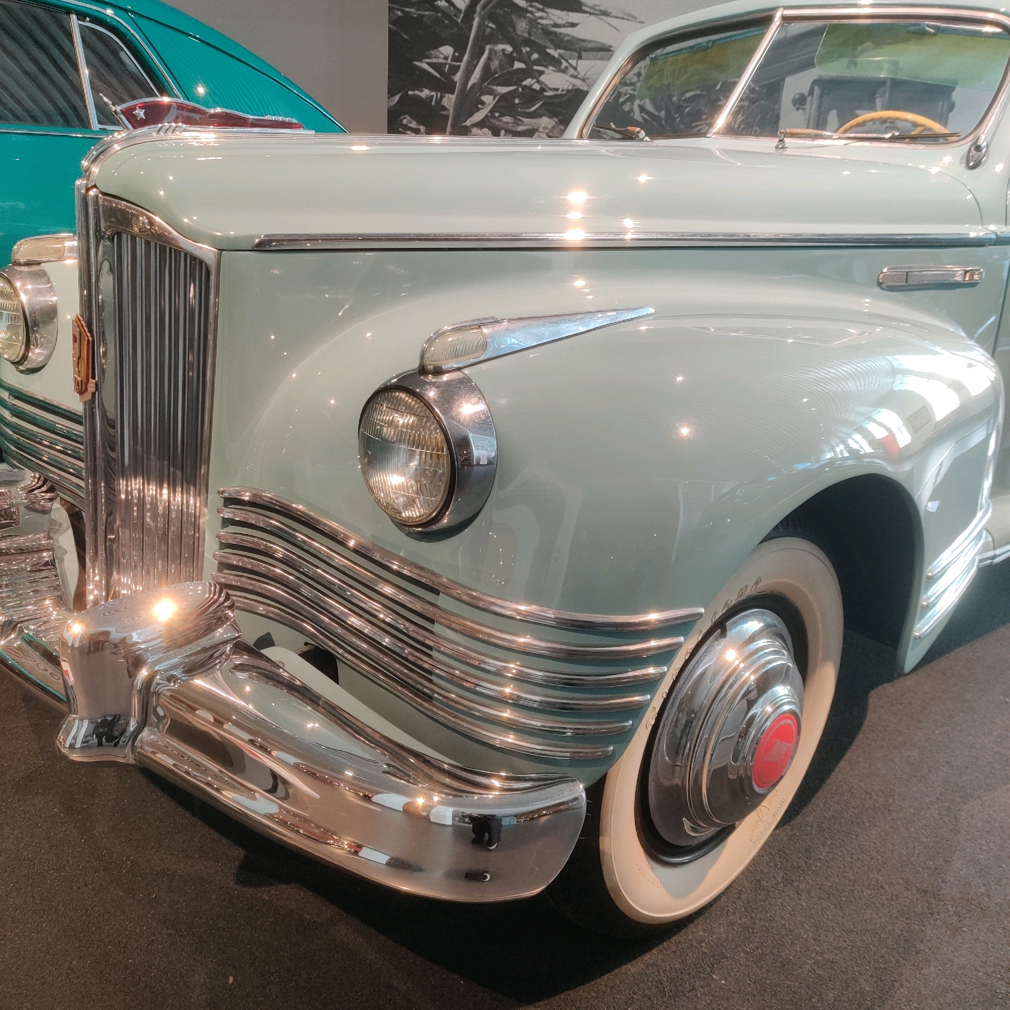 The image shows a close-up view of the front of a light blue vintage car, likely on display. The car features prominent chrome accents, including a vertical grille, a rounded bumper, and horizontal embellishments along the side. A circular headlight with a chrome bezel is visible on the left side. The wheel is also visible, with a whitewall tire and a chrome hubcap featuring a red emblem. In the background, the rear section of a teal-colored vintage car is partially visible. The setting appears to be an indoor exhibition space, possibly a museum, as indicated by the dark flooring and the lighting that creates reflections on the car's polished surfaces. This scene is located within the "Tank Museum" (Ural Military History Museum) in Verkhnyaya Pyshma, Russia, which houses a significant collection of Soviet-era automobiles and military vehicles.