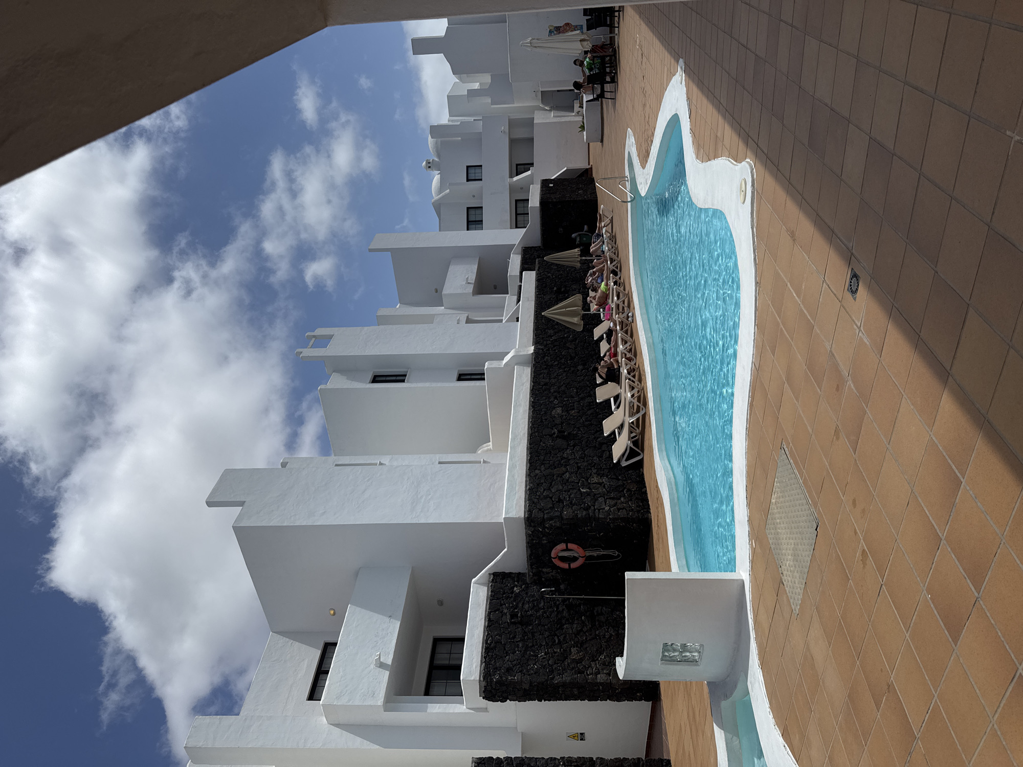 The image captures a view of a hotel or apartment complex in Costa Teguise, Spain. The architecture features a modern, white-washed style with tiered balconies and black volcanic rock accents. A swimming pool with clear blue water is adjacent to the building. Lounge chairs are positioned along the pool, occupied by several individuals. The surrounding area is tiled. A life preserver is visible on a wall. The sky is blue with scattered clouds. The scene appears to be taken during daylight hours.