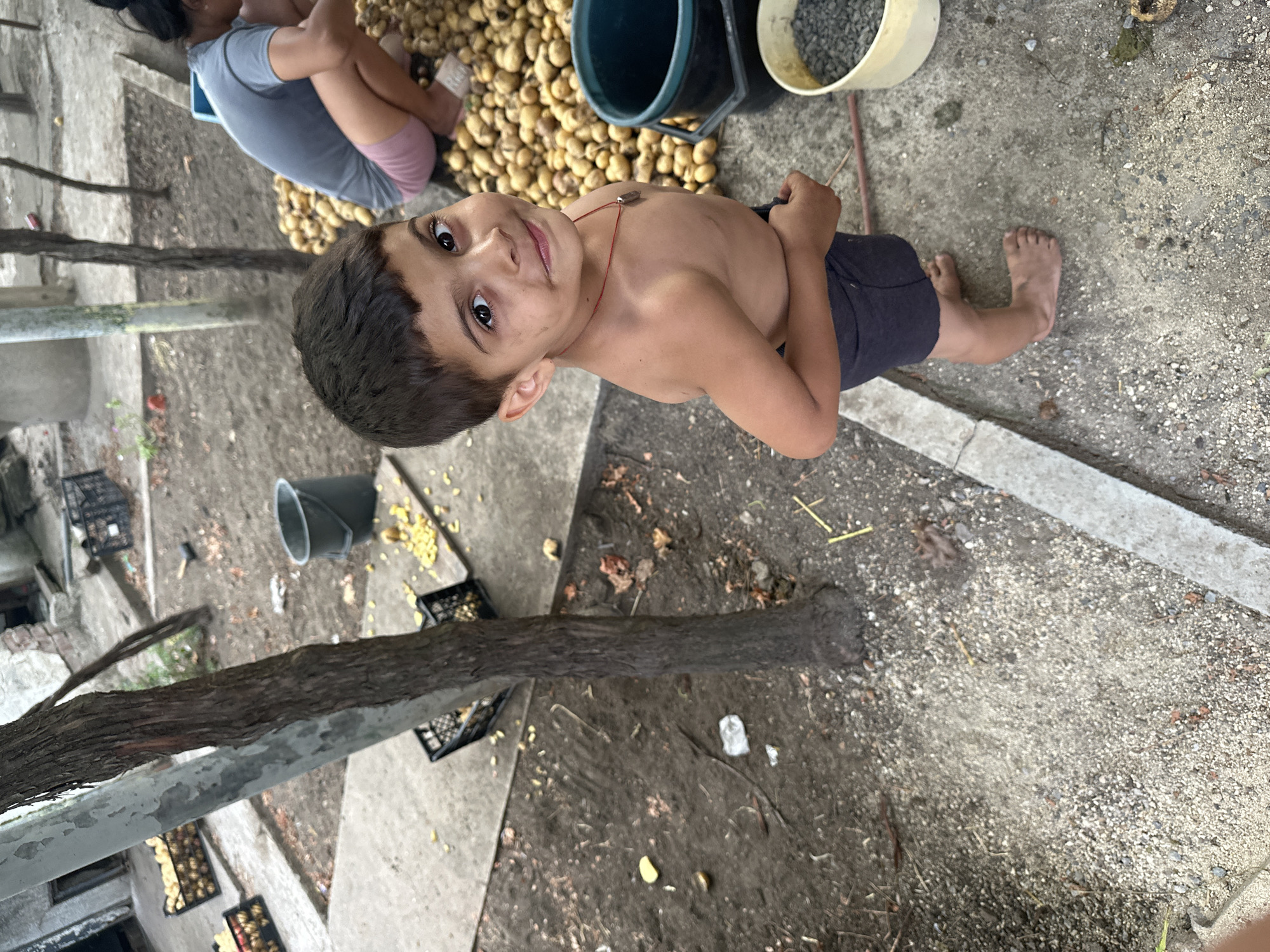 The image depicts a young boy, appearing to be around five to seven years old, standing barefoot on a dirt and concrete surface in what appears to be a rural or outdoor setting, potentially in Mlekarevo, Bulgaria. The boy is shirtless, wearing dark shorts, and looking directly at the camera with a slight smile. He has dark hair and brown eyes and is wearing a red cord necklace with a small pendant.
Behind the boy, to the right, a large pile of potatoes is visible, and a woman in a grey t-shirt and pink shorts is crouching, appearing to sort or handle the potatoes. Two buckets are also present; one dark blue or black, and a lighter colored one filled with small grey stones or gravel.
In the background, there are more crates of what appear to be potatoes, as well as some scattered debris and foliage. A tree trunk is prominent in the left and center of the frame. The overall environment suggests an agricultural or outdoor work area, possibly a backyard or a farm-related space. The lighting indicates it is daytime, with shadows present.