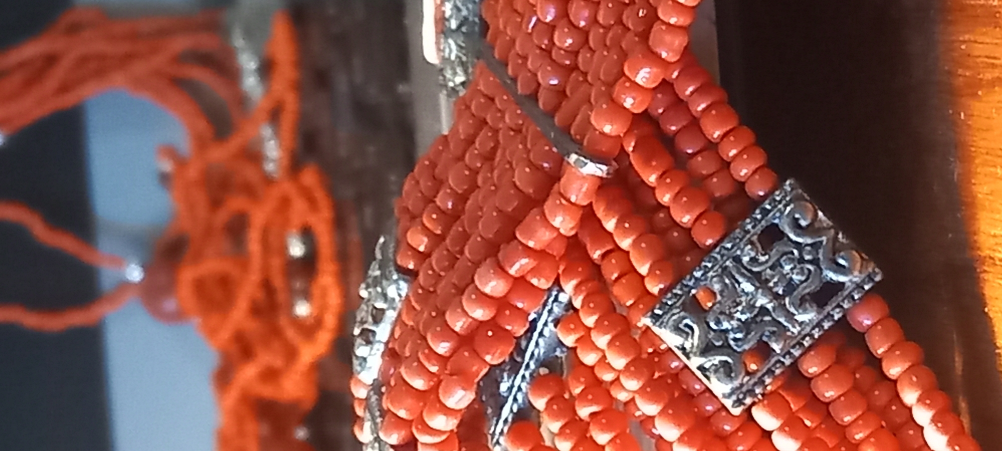 The image displays a close-up, angled view of a piece of jewelry, likely a multi-strand necklace or bracelet, made predominantly of small, round, orange-red beads. The beads possess a smooth, somewhat glossy finish and a consistent, vibrant hue, resembling polished coral.
Several ornate, silver-colored metal components are integrated throughout the beaded strands. A prominent rectangular metal piece in the foreground features an intricate, filigree-like design with swirling patterns, providing a decorative contrast to the natural beads. Smaller, plainer silver rings are also visible, holding together groups of bead strands.
The jewelry is sharply in focus on the right side of the frame, highlighting the texture of the beads and the detailed craftsmanship of the metalwork. The background is heavily blurred, but indistinct shapes of more orange-red beaded strands can be seen, along with areas of light blue and darker tones, possibly indicating other items on display or ambient reflections.
The image is a static shot of the jewelry; no people or specific activities are depicted. Details such as time of day, weather, or particular emotions are not discernible. No visible text is present within the image. The general setting appears to be indoors, possibly a display area for jewelry in Kryvyi Rih, Ukraine.