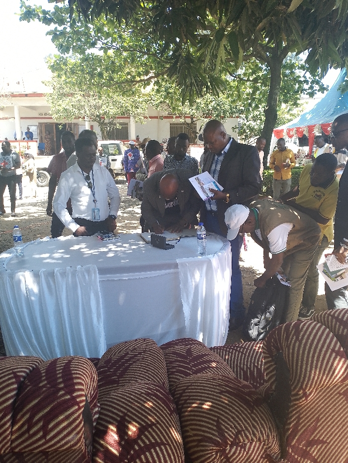 The image depicts a group of men gathered around a white table draped in a tablecloth. The men are in the process of signing a document. There is a black briefcase and two bottles of water on the table. The men are standing in front of a building. There are multiple vehicles parked in the background. The image was taken in Bunia, Democratic Republic of the Congo, at the coordinates 1.557 latitude and 30.244 longitude.