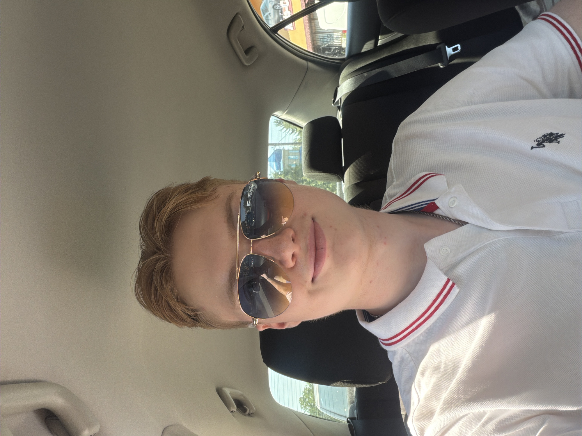 The image shows a young male with light brown hair wearing aviator sunglasses and a white polo shirt with red and blue trim on the collar and sleeves. He is seated in the back seat of a car, as indicated by the seatbelt, car interior, and windows. The windows show a blurred background of a city street with buildings and trees, suggesting an urban environment. The car appears to be in motion. One of the windows reflects a building with a blue dome, which could be a church or religious structure. The other window shows a commercial building with signage that includes the Cyrillic word "Авто". There is no specific information in the image that would allow for precise identification of the location within Polonne, Ukraine, beyond the general context of an urban setting.