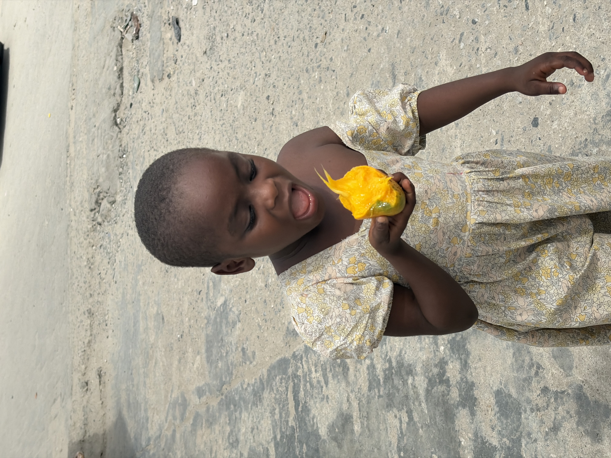 A young, dark-skinned child in a floral-patterned dress stands outdoors on a concrete or paved surface. The child is holding a bright yellow fruit, possibly a mango, and appears to be taking a bite or about to eat it, with their mouth open. The background suggests a street or outdoor area in Igboefon, Nigeria. The overall impression is of a candid snapshot of everyday life.