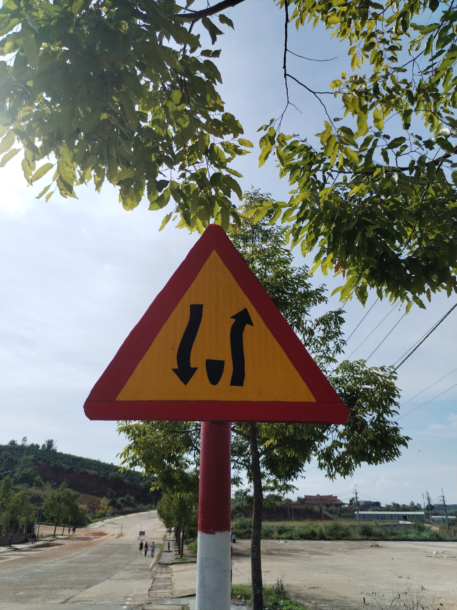 The image captures a "Two-Way Traffic Ahead" sign on a road in Bo Y, Vietnam. The sign is triangular with a red border and yellow background, displaying black arrows indicating opposing directions of traffic. The signpost is red and white. The road is asphalt with visible wear. A group of people are walking in the distance along the road. Trees flank the roadside. The sky is lightly clouded. In the background, there is a grassy field and buildings. A forested hillside is seen in the far left of the background.