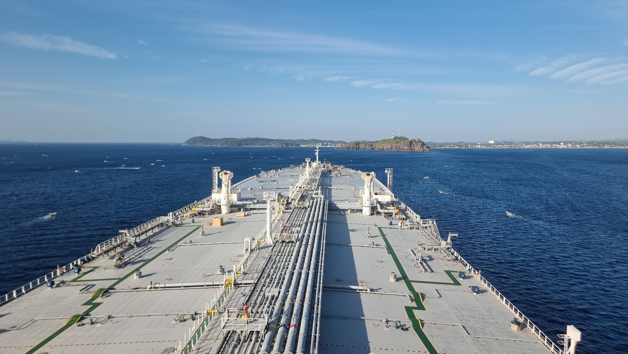 This is a wide-angle, high-angle, outdoor, daytime shot of the deck of a large cargo ship at sea. The ship is moving forward and the camera is positioned at the stern, looking towards the bow. The deck is gray and features numerous pipes, walkways, and safety equipment. In the distance, there is a shoreline with a city and hills. The sea is a deep blue and there are several small boats visible on the water. The sky is clear with a few wispy clouds. The scene is calm and peaceful, with no people or animals visible on the ship. The location in the city of Trincomalee, Sri Lanka, cannot be determined from the image alone.
