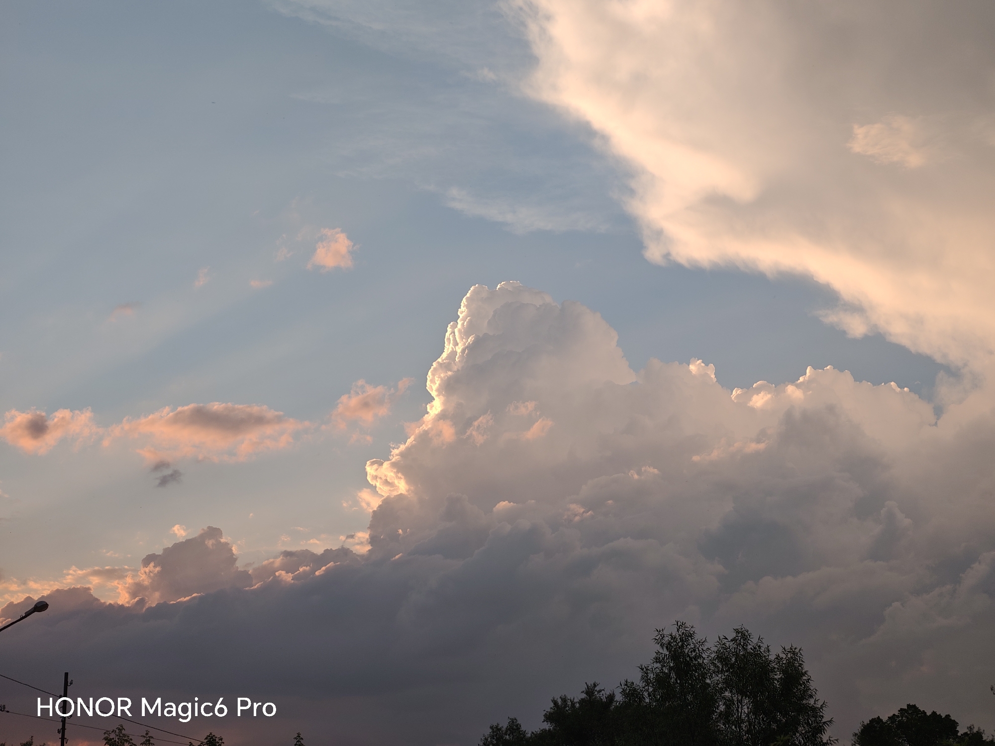 The image depicts a sky filled with dramatic clouds during what appears to be either sunrise or sunset. The sky is a gradient of blue and pink hues, with the sun's light illuminating the edges of the clouds, creating a luminous effect. Several layers of clouds are visible, with some appearing dark and heavy at the bottom, and others light and wispy near the top. The text "HONOR Magic6 Pro" is overlaid in white in the lower-left corner of the image. In the lower portion of the frame, silhouetted trees and a street lamp are visible against the sky, suggesting an outdoor, possibly urban or suburban setting. The scene is static, with no discernible human activity or interaction. The weather appears overcast with dramatic cloud formations. The location is identified as Turbiv, Ukraine.