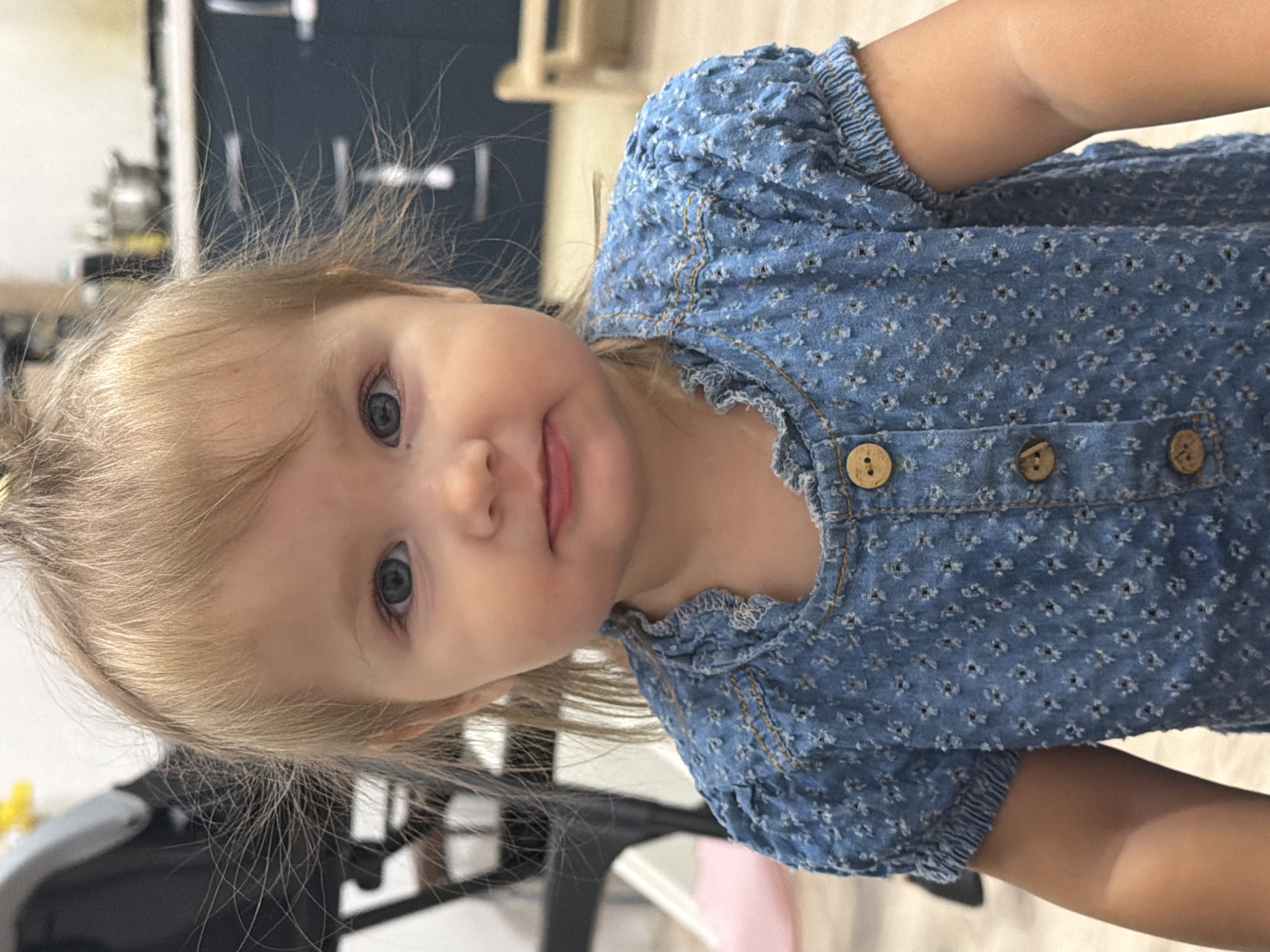 A medium shot captures a young Caucasian child with blonde hair and blue eyes, looking directly at the camera. The child is wearing a blue denim-like dress with a floral pattern and wooden buttons. The background is blurred, showing elements of a home interior, including kitchen cabinets and furniture. The lighting is natural and appears to be indoors. There is no visible indication of the child's specific location within the city of Krasnyi Krym, Russia.