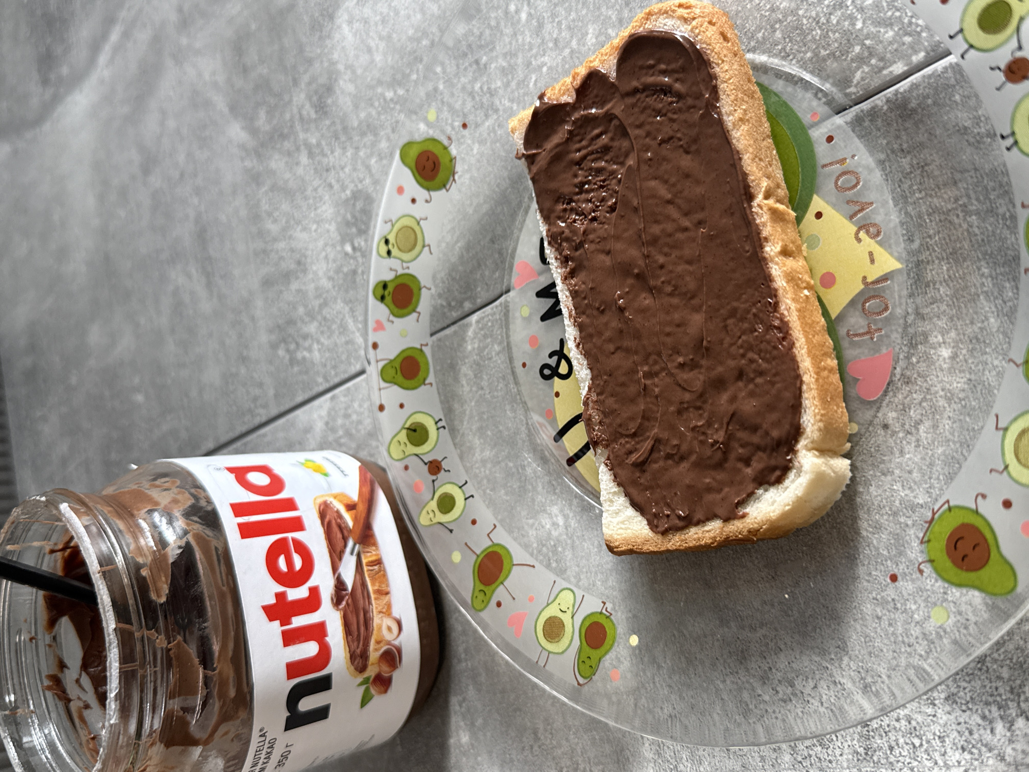 An overhead shot displays a simple snack arrangement on a gray, textured surface. The central object is a slice of bread generously spread with chocolate hazelnut paste, resting on a clear plastic plate. To the left, a glass jar of Nutella, partially consumed, stands open with a dark utensil (likely a spoon or spatula) inserted into the spread.
The bread appears to be a slice of white bread, lightly toasted, and thickly coated with the rich, dark brown Nutella, showing visible streaks from its application. The plate is clear plastic with a decorative edge, featuring an array of cartoon avocados in various joyful poses, some smiling, some with heart-shaped cheeks. Scattered across the plate are small colorful dots and delicate pink heart shapes. Partially visible and inverted, the text "I love Jo-Yo!" or "I ♥ Jo-Yo!" is inscribed on the plate.
The Nutella jar's label prominently displays the brand name "Nutella" in red lettering. Below it, smaller text includes "NOIX DE CAKAO" and "350 г" (350 grams). The label also depicts Nutella being spread on bread, alongside hazelnuts. The contents of the jar show the remaining dark brown spread.
The background is a consistent gray, suggesting a countertop or table. The scene depicts a casual moment, likely the preparation or enjoyment of a snack or light meal. No people are visible in the image.