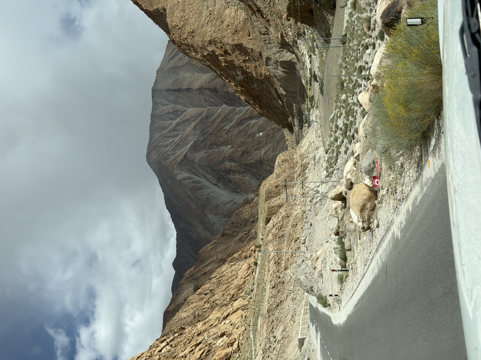 The image depicts a mountainous landscape with a winding road. The mountains are steep and rocky, with varying shades of brown and gray. The road is paved and appears to be a two-lane highway, curving to the left and disappearing around a bend. The surrounding terrain is arid and sparsely vegetated, with scattered rocks and low-lying bushes. Overhead, the sky is partly cloudy, with patches of blue visible between the white and gray clouds. The overall impression is one of a remote and rugged natural environment. The location is identified as being in the city of Hymia, India.