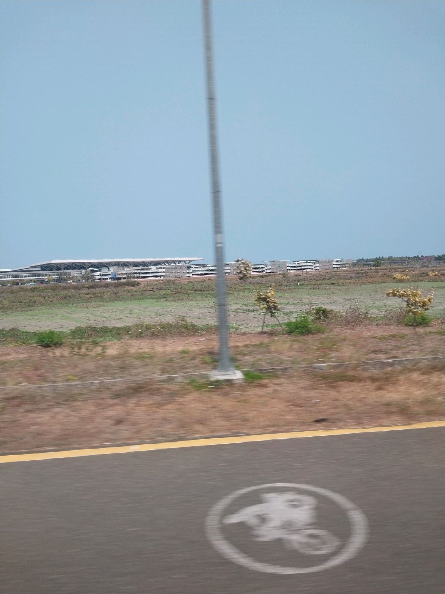 The image, captured on an overcast day, showcases a modern building complex in Glagah, Indonesia. The complex, with its distinctive cantilevered roof, stretches horizontally across the frame, suggesting a large public facility like an airport terminal or convention center. In the foreground, a paved road with a yellow line marking the edge is visible. A blurred bicycle lane symbol is painted on the asphalt, indicating a designated area for cyclists. The environment surrounding the building is a mix of dry, brown vegetation and patches of green, hinting at a semi-arid or seasonal landscape. A tall, slender pole, possibly a lamppost or communication tower, stands vertically in the center of the image, partially obscuring the view of the sky. The overall impression is one of a developing urban or semi-urban area, with modern infrastructure set against a natural, somewhat sparse backdrop.