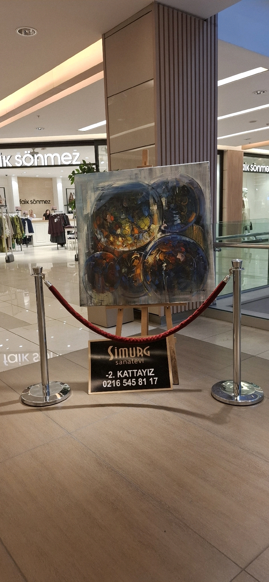 The image depicts an abstract painting displayed on an easel in what appears to be a shopping mall. The painting features circular shapes in shades of blue, orange, yellow, and black, with thick brushstrokes. In front of the painting, a black sign with white text is visible, displaying "Simurg Sanatevi" and contact information: "-2. KATTAYIZ 0216 545 81 17". This indicates the location is likely a gallery or art studio in Kadıköy, Turkey, given the Turkish text and the phone number format associated with Istanbul. The painting is roped off with stanchions and red velvet ropes, suggesting it is part of an exhibition or for sale. In the background, storefronts of clothing shops, including one labeled "Faik Sönmez", are visible, confirming the mall environment. The floor is tiled with a wood-like pattern. No individuals are directly interacting with the painting or the sign.