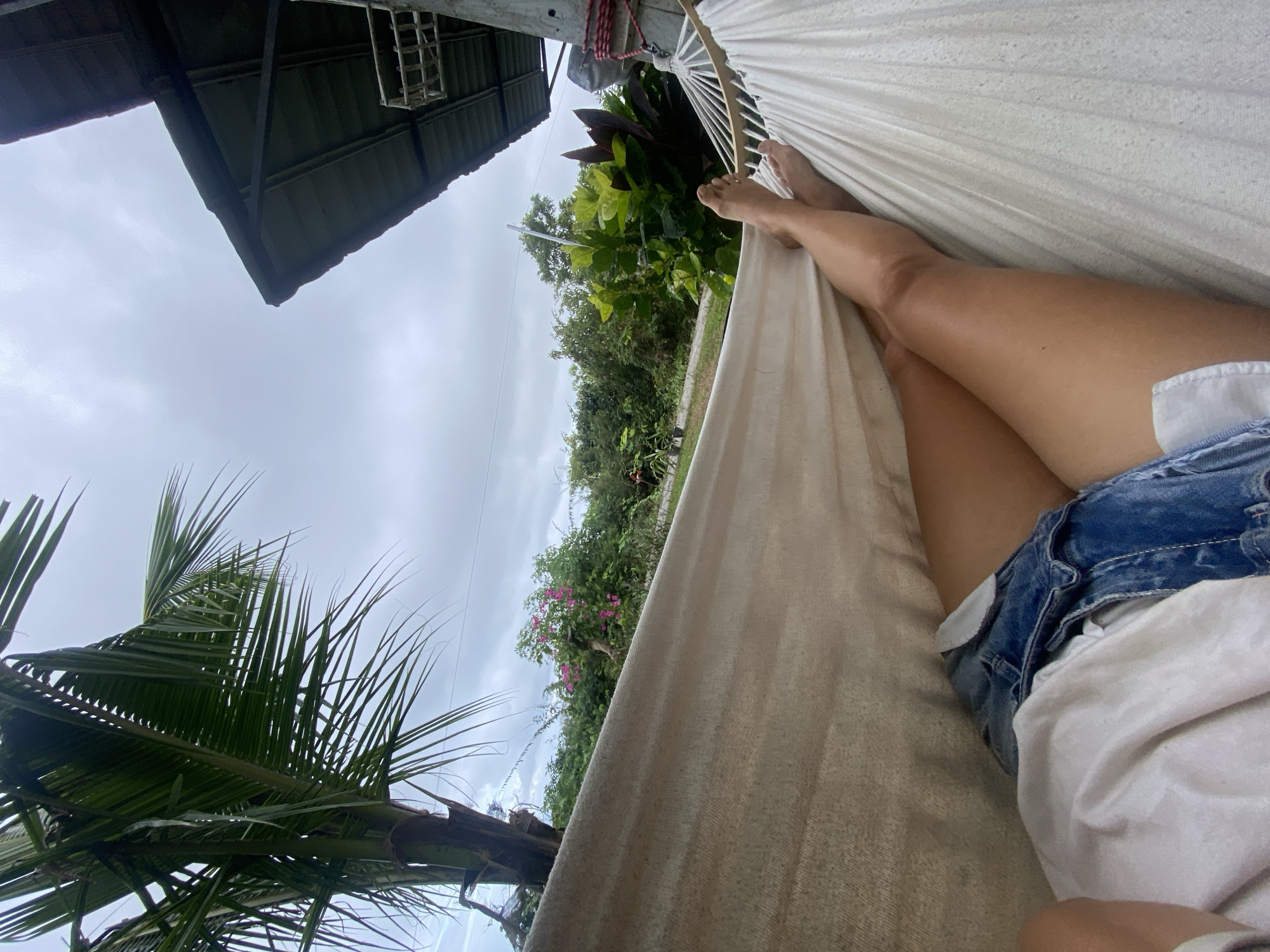 A person lies in a white hammock, their legs crossed and visible from the thigh down. They are wearing denim shorts and a white t-shirt. The hammock is suspended outdoors, with lush green foliage and flowering bushes in the background. A coconut palm tree is visible on the left, and a section of a building's roof overhang is on the right. The sky is overcast and cloudy. The scene appears to be in a tropical environment, possibly Kuta Selatan, Indonesia, given the presence of tropical vegetation.