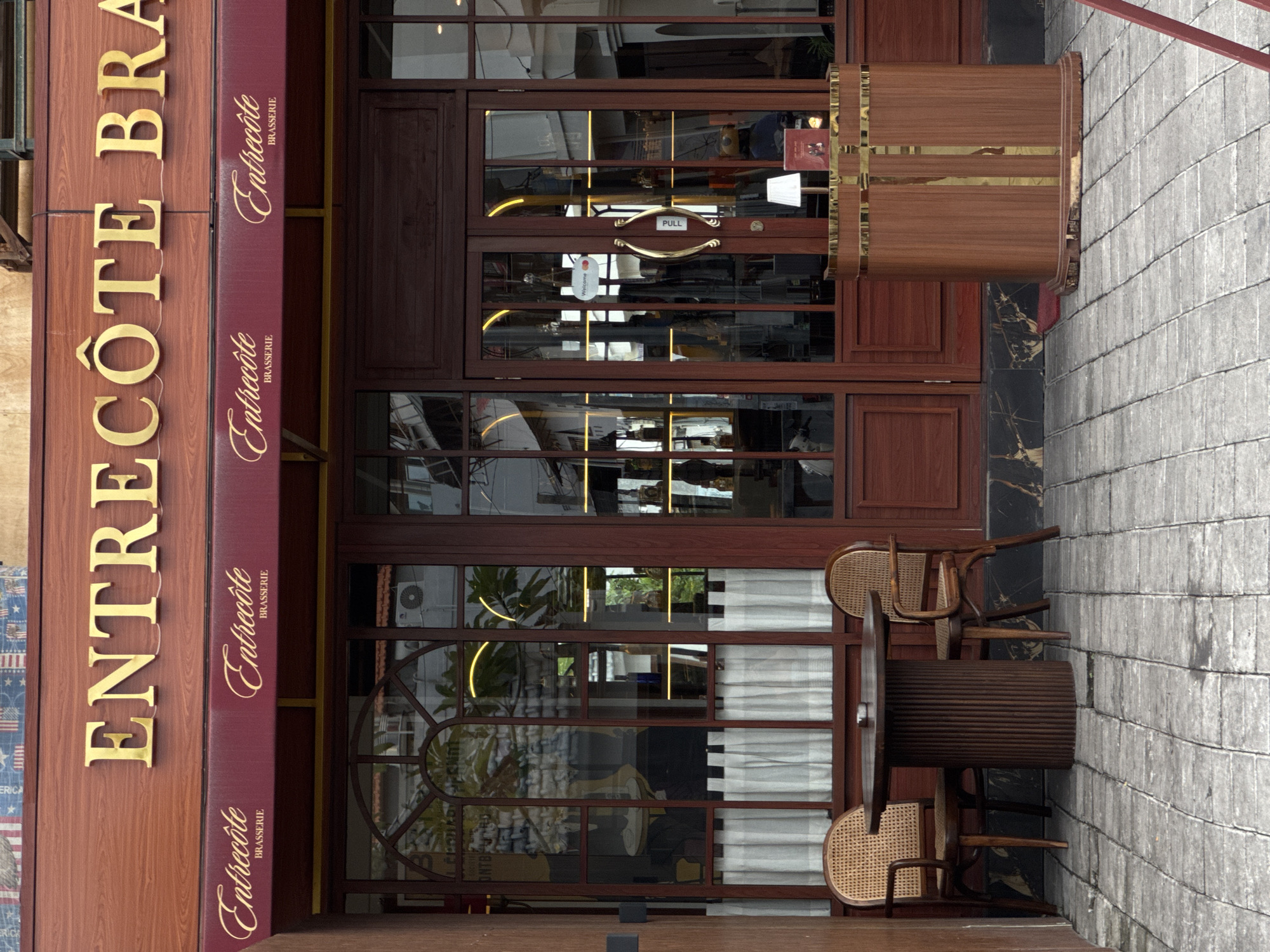 The image displays the exterior of a restaurant named "Entrecôte Brasserie" located in North Kuta, Indonesia. The building features a facade of dark wood panelling and extensive glass windows and doors.
Prominently displayed in large, gold, three-dimensional letters on the upper part of the facade is "ENTRECÔTE BRA," with "BRASSERIE" likely continuing off-frame. Below this, a maroon-colored awning also features "Entrecôte BRASSERIE" in elegant gold script.
The main entrance consists of large, wooden-framed glass doors. One of the door handles has a small, white rectangular tag with "Welcome" written on it and a black "PULL" sign. The reflections in the glass reveal parts of the interior, including yellow strip lighting and some indoor furnishings.
To the right of the main entrance, a tall, wooden, barrel-shaped structure with horizontal gold bands stands. Further to the right and slightly in front of the building, there is an outdoor seating area with two wooden chairs featuring woven cane backs and seats, and a large, round wooden table resembling a spool. The chairs and table are placed on a paved area, which appears to be a sidewalk or patio. A grey brick wall is visible on the far right, next to the outdoor seating.
The scene appears to be taken during the daytime, though the exact weather is unclear due to reflections. There are no people visible in the image, suggesting the restaurant might be closed or empty.