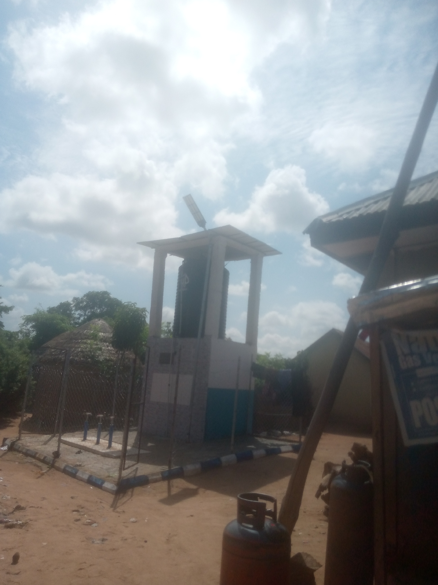 The image depicts an outdoor scene in Jalingo, Nigeria, featuring a water dispensing station. The central element is a tall, square structure with white pillars supporting a flat roof, housing a large black water tank. The base of the structure is tiled with a mosaic of grey and blue squares. In front of this structure, a fenced area contains three blue and silver water taps. To the left of the water station, a thatched-roof hut with a fence is visible. In the foreground and to the right, there are propane gas cylinders, one of which is red, and part of a building with a corrugated metal roof. A sign that reads "NPA" and "GAS" is partially visible on the right side of the frame. The ground is sandy and uneven, with scattered debris. The sky is partially cloudy with bright sunlight.