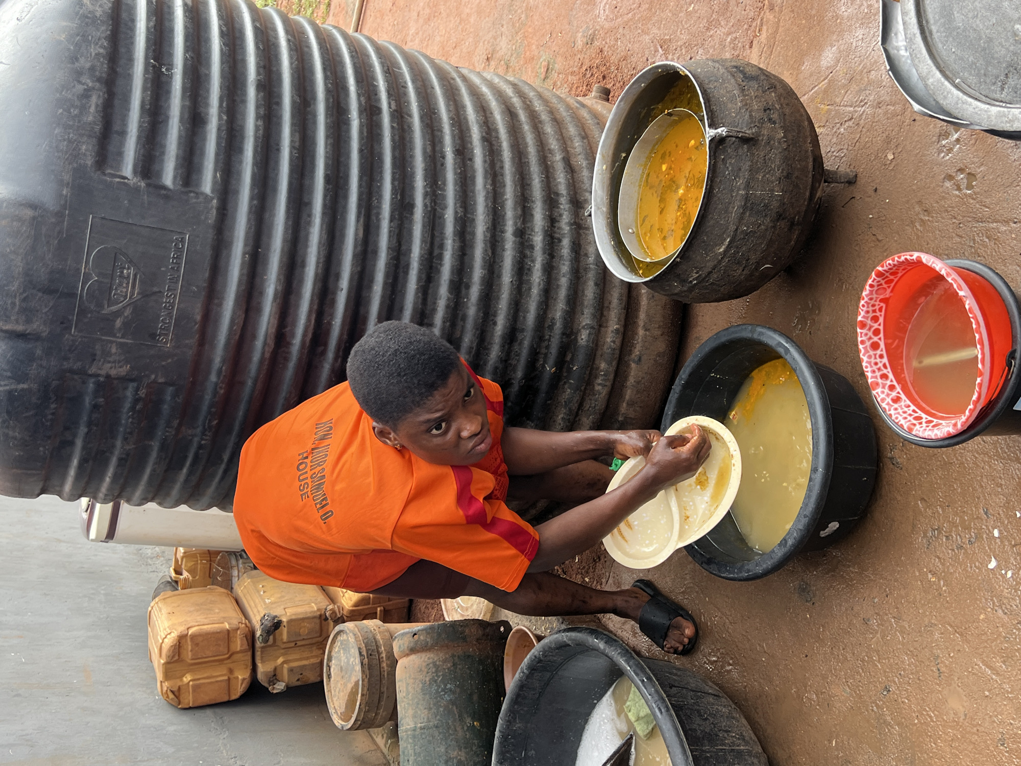 In Nkpor, Nigeria, a young person wearing an orange shirt with red accents and dark shorts is leaning over to wash a white plate with divisions. They are barefoot, wearing black flip-flops, and their hands are submerged in soapy water within a black plastic basin. Another black basin to the left contains more soapy water and a green sponge. In front of the person, a large, dark cauldron sits on a stand, holding a yellowish stew. To the right, a red bucket with a white patterned rim is filled with a brownish liquid. Behind them, a large, ribbed black water tank is visible, with a logo that reads "ONITSHA" and "STRONGEST IN AFRICA." To the left of the tank are several stacked brown plastic containers. The ground is wet and appears to be made of red earth, with some puddles reflecting the scene. The overall environment suggests an outdoor, utilitarian setting, possibly a communal cooking or washing area.