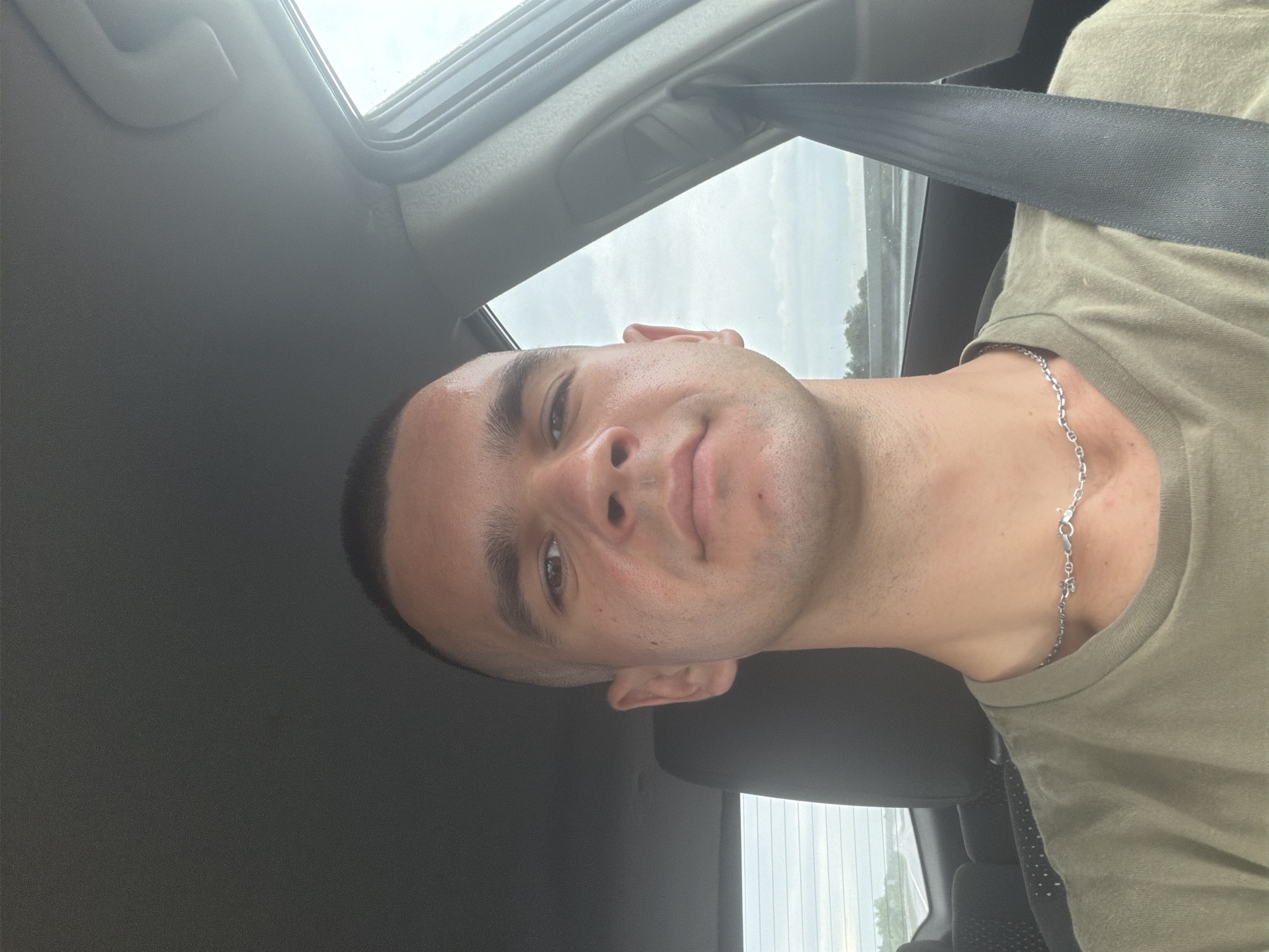 A young man with a buzz cut is taking a selfie from the driver's seat of a car. He is wearing a military green t-shirt and a silver chain necklace. The car's interior is visible, including the dashboard, steering wheel, and a seatbelt. The background shows a cloudy sky and a road, suggesting the man is on a highway. The location is identified as Kaloyanovets, Bulgaria.
