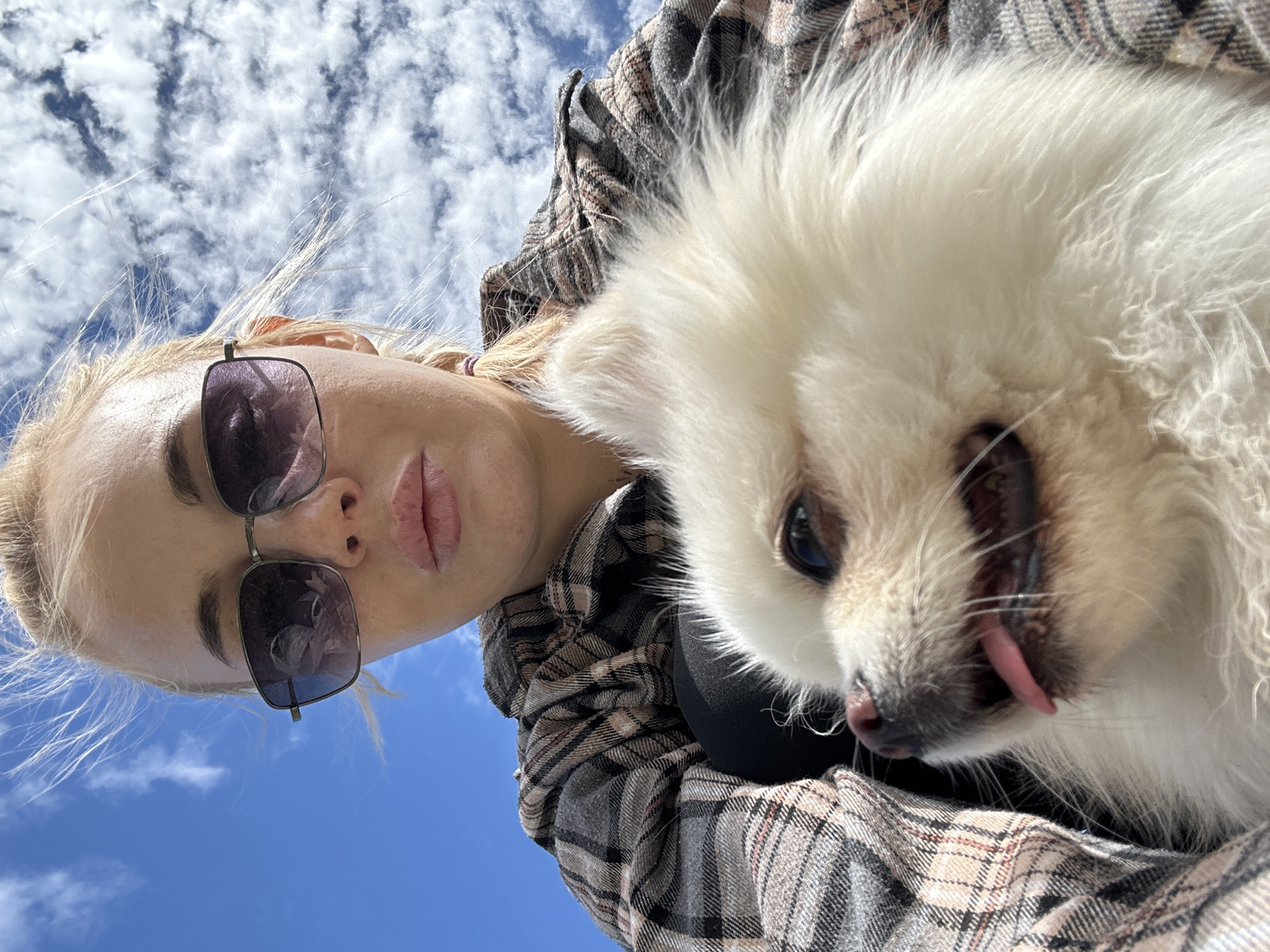 A selfie taken by a fair-skinned woman with blonde hair, wearing sunglasses and a plaid shirt. She is holding a small, white, fluffy dog, likely a Pomeranian, with its tongue slightly sticking out. The background shows a blue sky with scattered clouds. The location is possibly Высокий, Russia.