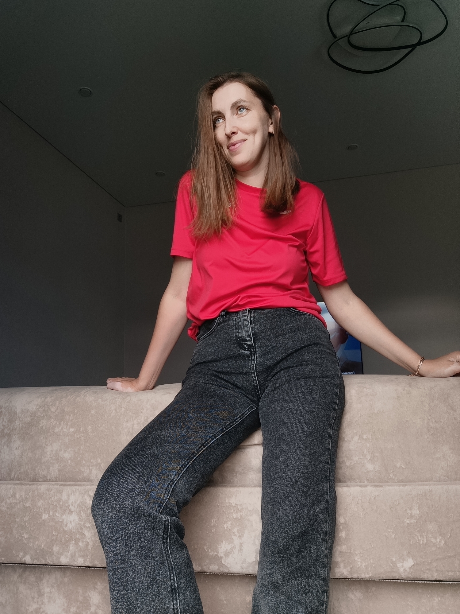 A woman with shoulder-length brown hair, wearing a red t-shirt and dark jeans, sits on a light-colored upholstered surface. She is leaning back and looking upwards and to her right with a slight smile. Her left arm is extended and resting on the surface, while her right arm is bent at the elbow and also resting on the surface. Her legs are crossed. The background is a dark, dimly lit room with a dark ceiling. A modern ceiling light fixture with a circular design is visible on the ceiling. The scene is indoors, likely in a residential setting, and there is no indication of the specific location in Chaikovsky, Russia, beyond it being an interior.
