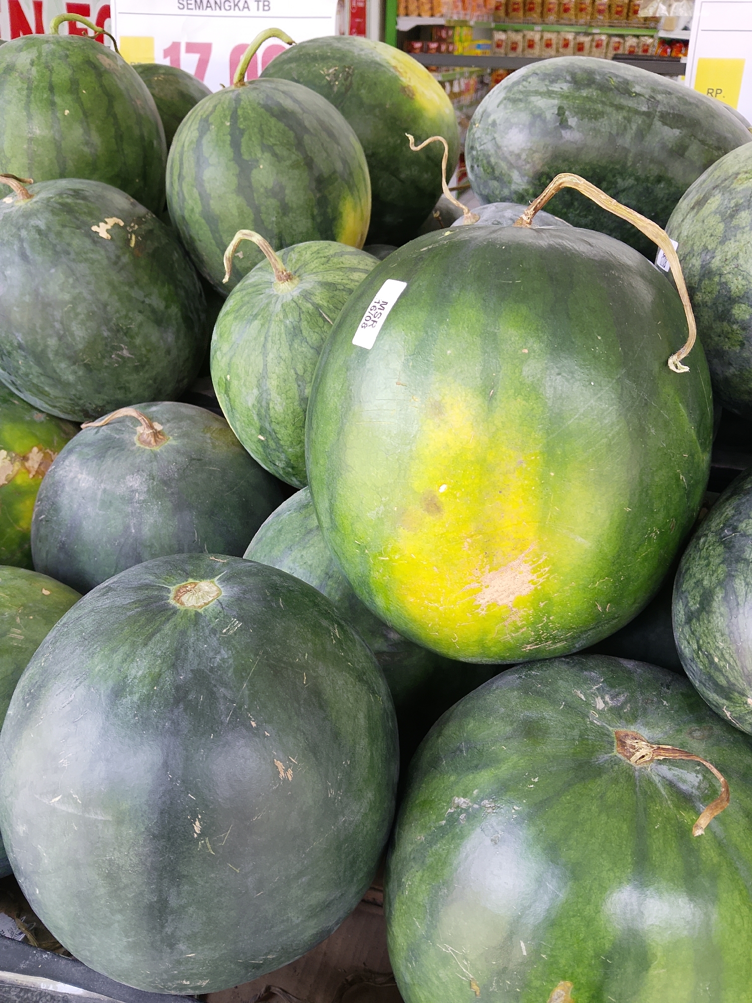 The image shows a close-up view of a pile of watermelons, likely at a market or roadside stall. The watermelons are mostly dark green with some lighter green stripes and yellow patches on their surfaces. Some watermelons have their stems attached. In the background, there are shelves stocked with various products, possibly food items, and a sign that reads "SEMANGKA TB". The overall scene suggests a commercial setting where watermelons are being sold. The location is identified as Karangboyo, Indonesia.