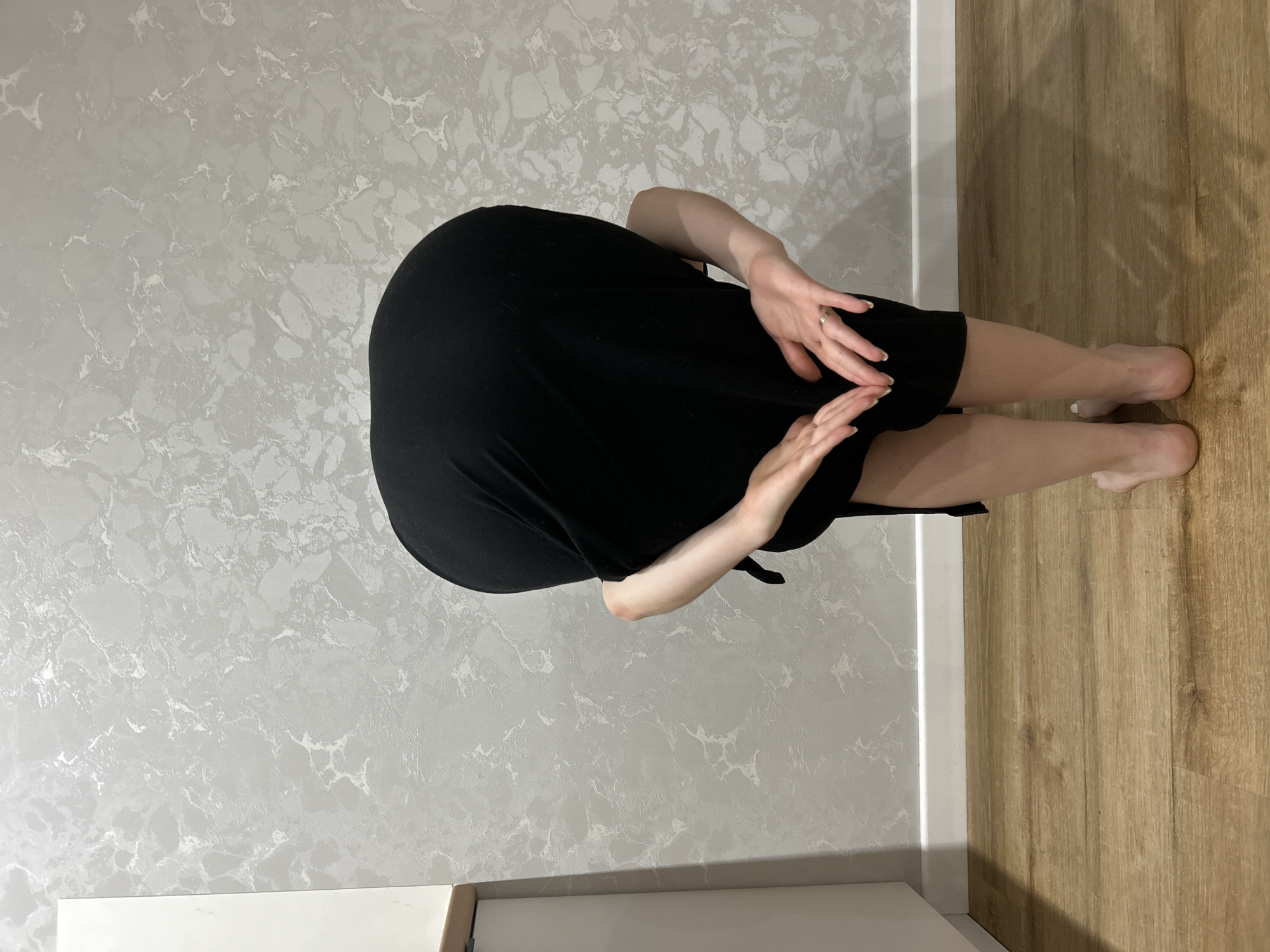A person wearing a black dress is standing in a room. The room has gray marble-patterned walls and light brown wood floors. The location is unknown, but potentially in Chaikovsky, Russia. The person's hands are resting on their dress.