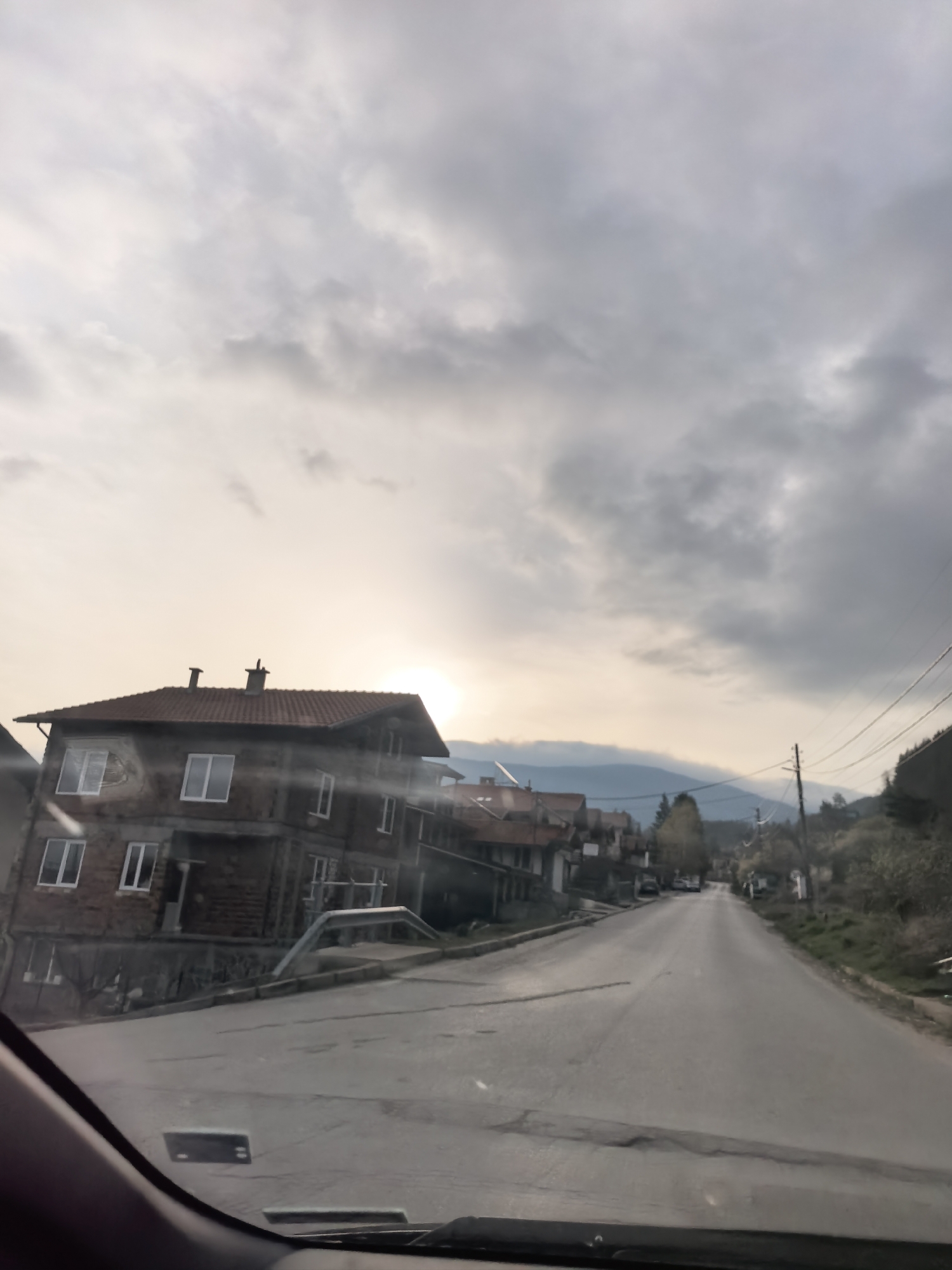 The image shows a street in a town under a cloudy sky. The sun is partially obscured by clouds, creating a diffused light. On the left side of the road, there is a two-story building with a red tile roof and white-framed windows. The building appears to be under construction or renovation, with some unfinished brickwork. The road itself is paved and has some cracks and patches. It leads uphill into the distance, with other buildings and trees visible along the sides. On the right side of the road, there is vegetation, including trees and bushes. Power lines run along the right side of the road. The photograph appears to have been taken from inside a vehicle, as part of the dashboard and windshield are visible at the bottom of the frame.