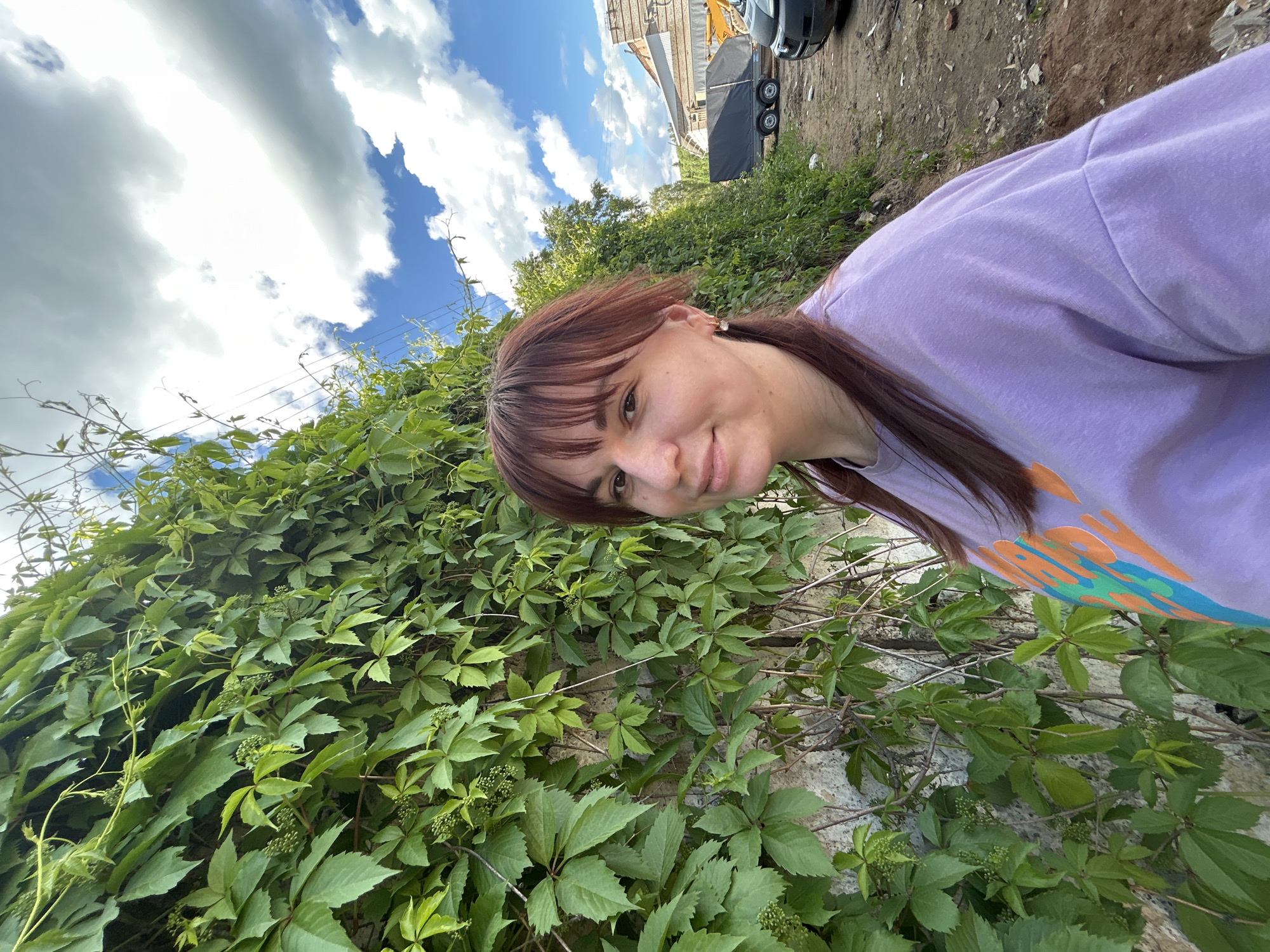 A woman with reddish-brown hair and bangs is shown from the upper torso up in a self-portrait style photograph, with a background featuring dense green foliage on a rough, light-colored wall. The sky is partially visible in the upper left, showing a mix of blue and white clouds. Above the foliage and wall, glimpses of a construction site or industrial area are discernible, including equipment such as a car, trailer, and construction vehicle. The woman is wearing a lavender-colored shirt with an orange and blue design on the front. The location is implied to be Chaikovsky, Russia. The photograph's orientation appears to be slightly tilted.