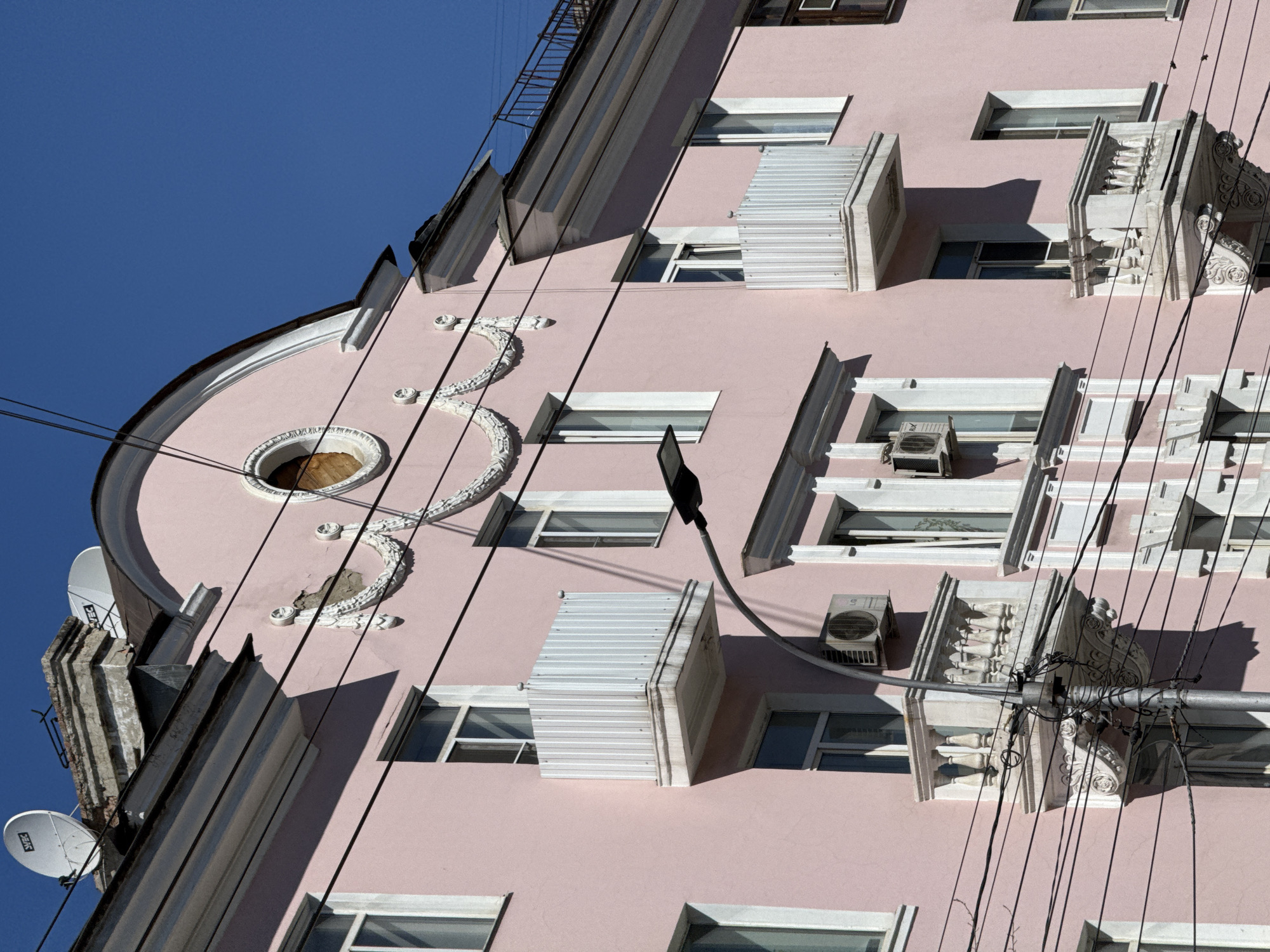 An eye-level, medium shot shows a portion of a pink residential building in Chita, Russia. The building is angled, appearing as if viewed from the corner of the structure. Several white-framed windows are visible, some with metal awnings above them. A white decorative element resembling a stylized floral pattern is present on the building's facade. Two satellite dishes are mounted on the left side of the building. Numerous electrical wires are strung across the front of the building. A street light fixture is mounted on the building's facade.