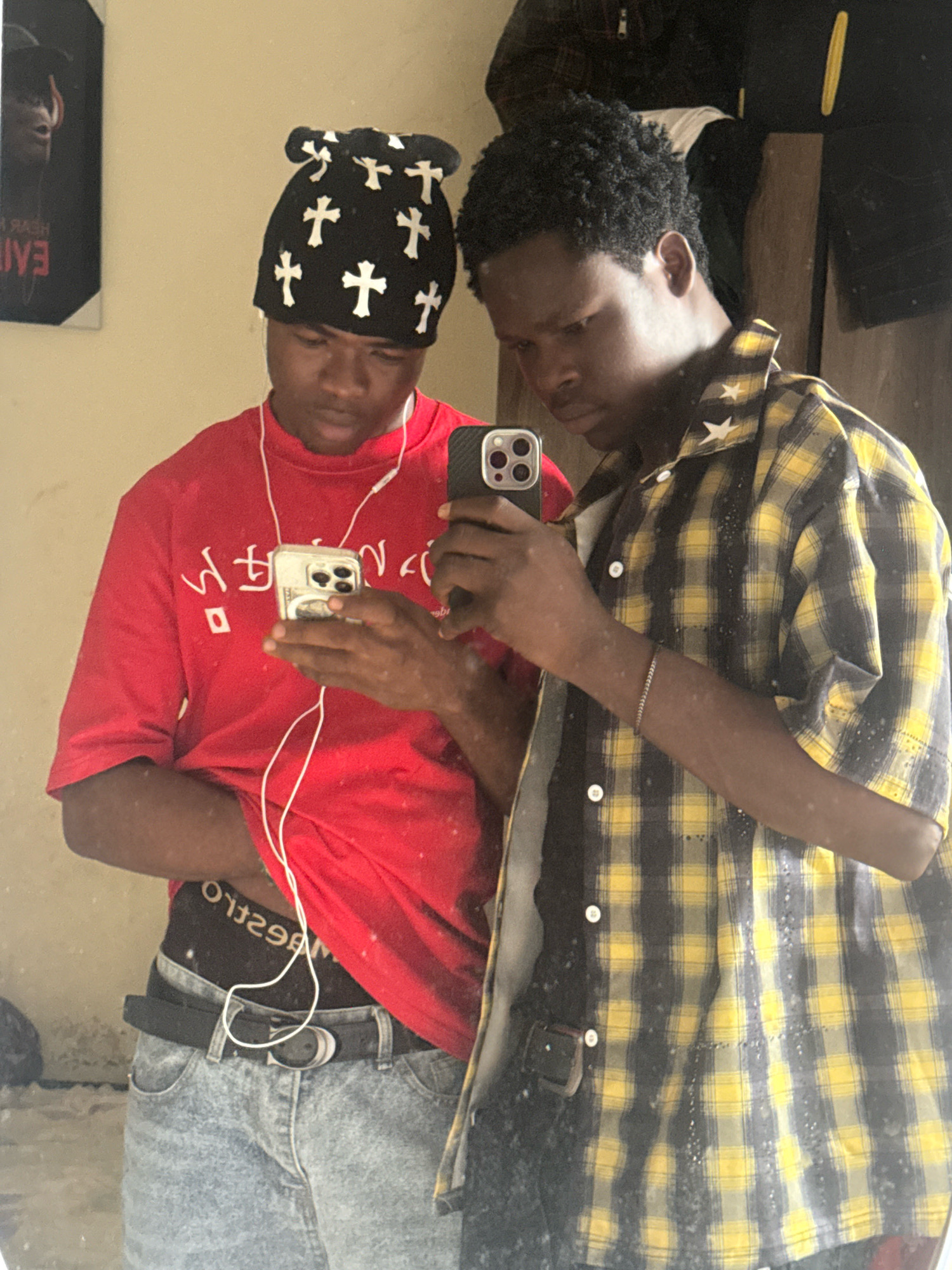 Two young men are reflected in a mirror, with the background appearing to be an indoor space in Ihiagwa, Nigeria. The man on the left is wearing a red t-shirt with white graphics and a black beanie adorned with white crosses. He is holding a smartphone. The man on the right is wearing a yellow and black plaid shirt with white stars and is also holding a smartphone. Both individuals are looking at their phones. The lighting in the room is somewhat dim, and there is a painting on the wall behind the man on the left. The environment suggests a casual setting, possibly a home.