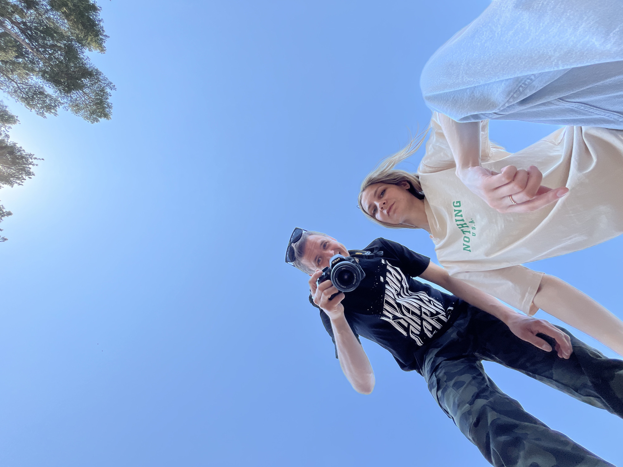 The image shows a man and a woman from a low angle. The man is holding a Nikon camera and wearing a black t-shirt with a white graphic, camouflage pants, and sunglasses. The woman is wearing a light-colored t-shirt with green lettering and light-wash jeans. The background is a clear blue sky with some trees visible in the top left corner. The location is Chaikovsky, Russia.