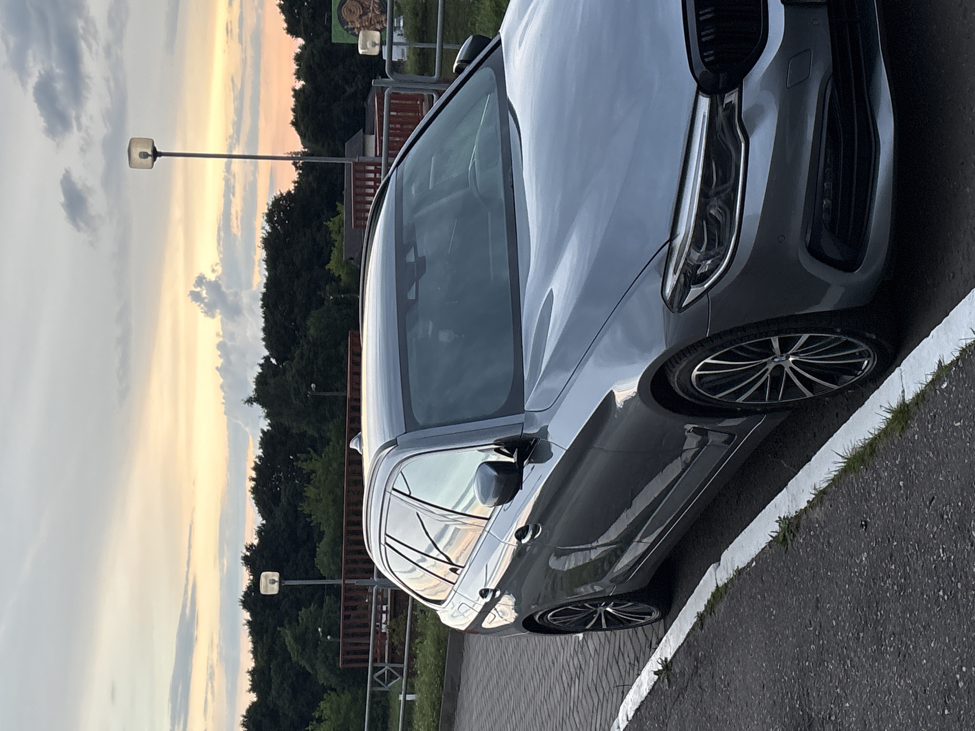 A gray station wagon is parked on a paved area next to an asphalt road. The car is facing the right side of the image, with its front angled slightly towards the camera. The sky above is a blend of pale orange, yellow, and gray clouds, suggesting it is either dawn or dusk. Behind the car, a line of dark green trees forms a dense backdrop. A red railing and a metal fence are visible in the mid-ground. There are two lampposts in the background, one on the left and one on the right, each with a square, frosted light fixture. The car has alloy wheels with a multi-spoke design. The scene appears to be an outdoor area, possibly a parking lot or the side of a road in a suburban or rural setting. The location is not specified as Vitsebsk, Belarus.