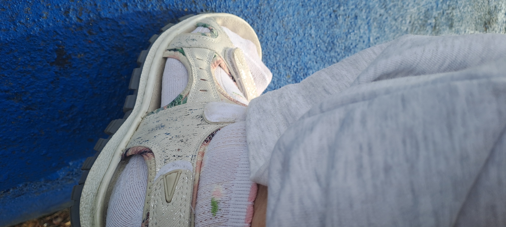 A close-up shot focuses on a person's foot wearing a white sandal with a floral pattern and white socks. The sandal is resting on a textured blue surface. A light grey garment is draped over the foot and sandal, partially obscuring the view. The scene appears to be outdoors, possibly in a park or playground, given the textured blue surface, which could be playground surfacing. There are no specific landmarks or architectural features visible to determine the exact location within Castlebar, Ireland. The lighting suggests daylight.