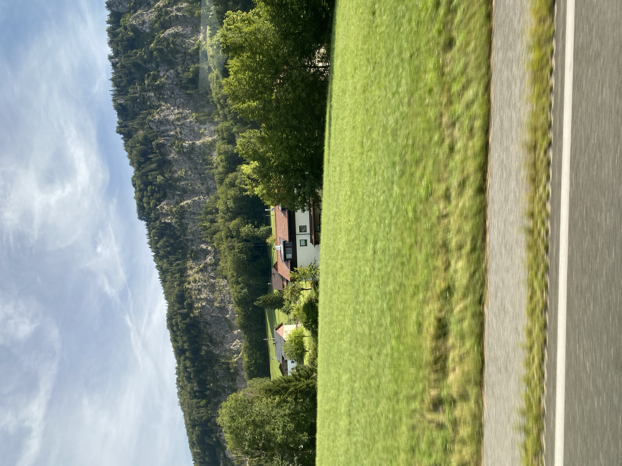 The image depicts a rural landscape in the vicinity of Winkl, Austria. In the foreground, a blurred green field extends across the frame, suggesting movement, possibly from a vehicle. A gray asphalt road with a white line runs horizontally across the bottom of the image, also exhibiting motion blur. In the mid-ground, several houses with red-tiled roofs are visible amidst lush green trees. These structures appear to be residential buildings, one of which has a prominent white facade and dark-trimmed windows. A steep, rocky mountainside covered in dense green forest dominates the background, reaching towards a pale blue sky with wispy clouds. The overall scene conveys a serene, natural environment characteristic of a mountainous region.