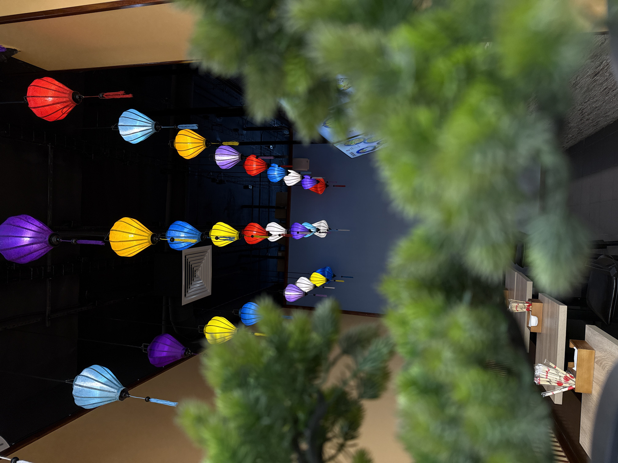 This is an indoor photograph depicting the interior of a commercial establishment, likely a restaurant or cafe, situated in Moscow, Russia. The scene is dominated by numerous colorful, illuminated lanterns suspended from a dark, industrial-style ceiling. The lanterns are of various balloon or teardrop shapes and sizes, glowing in shades of purple, yellow, blue, red, and white, with several featuring decorative tassels. The ceiling structure includes visible vents and dark beams. Walls are visible in the background, featuring light brown and darker grey-blue sections. The foreground is occupied by heavily blurred green foliage, consistent with an artificial decorative plant, which partially obscures the lower and left areas of the image. Through the foliage and beneath the hanging lanterns, multiple light-colored wooden tables are arranged in rows, each furnished with wooden containers holding chopsticks. A dark chair is partially visible under one table. In the distant background, a digital display screen, possibly presenting menu items with circular graphics, is faintly discernible. No people are present, and no actions are taking place.