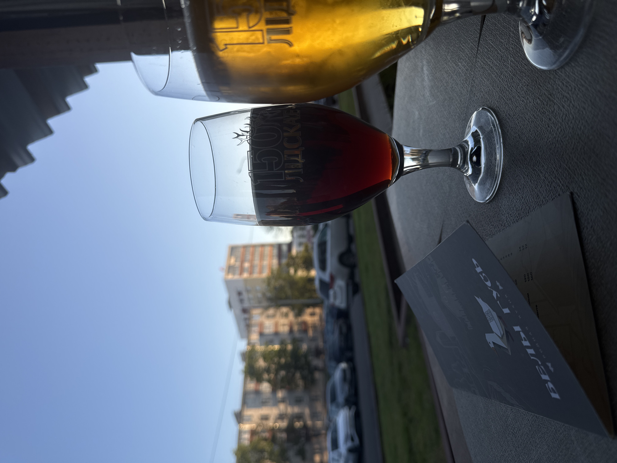 Two glasses of drinks are on a dark, textured surface, possibly a table. The glass in the foreground is filled with a dark red liquid, likely wine or a dark beer, and has text etched into it. The glass behind it contains a lighter, golden-colored liquid, possibly beer or white wine, and also has etched text. A black menu or booklet is placed on the surface near the glasses. In the background, a city scene is visible, with buildings and parked cars under a clear blue sky. The lighting suggests it is daytime, likely late afternoon or early evening, with the sun casting shadows. The overall scene appears to be an outdoor setting, possibly a cafe or restaurant patio. The text on the glasses and menu is in Russian. The text on the darker glass reads "МУРСК ЛЮДСКАЯ". The text on the menu is partially visible and appears to be a list of items with prices.